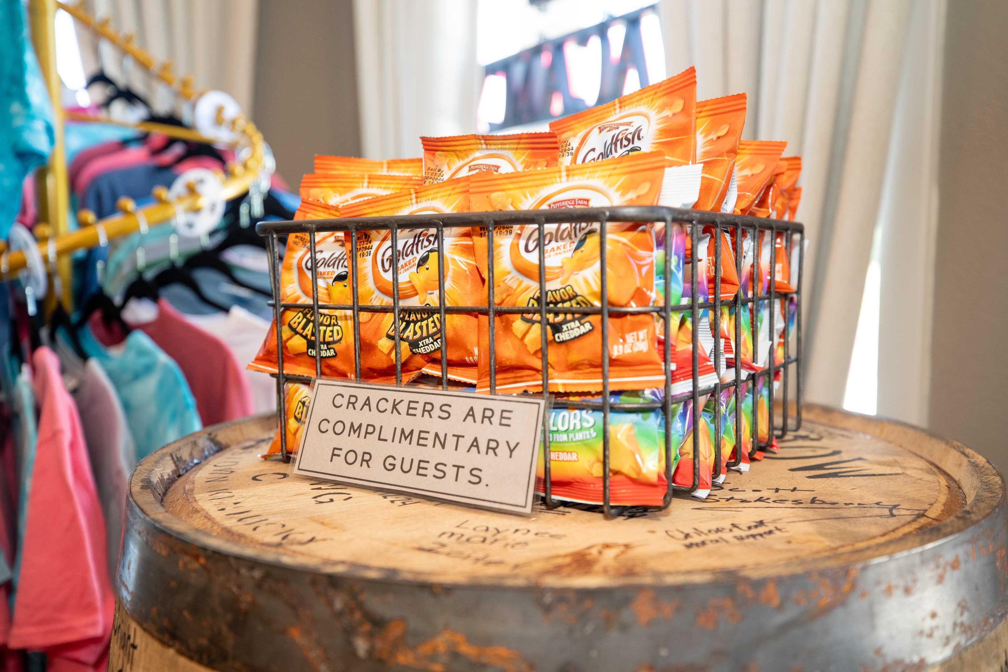 A wire basket filled with assorted packs of crackers labeled as complimentary for guests, placed on a wooden barrel.