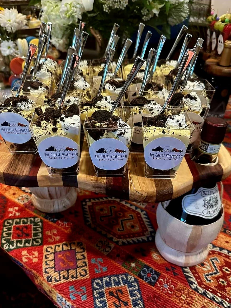 A wooden platter holds individual dessert cups topped with whipped cream, crushed cookies, and spoons, surrounded by a colorful patterned tablecloth.