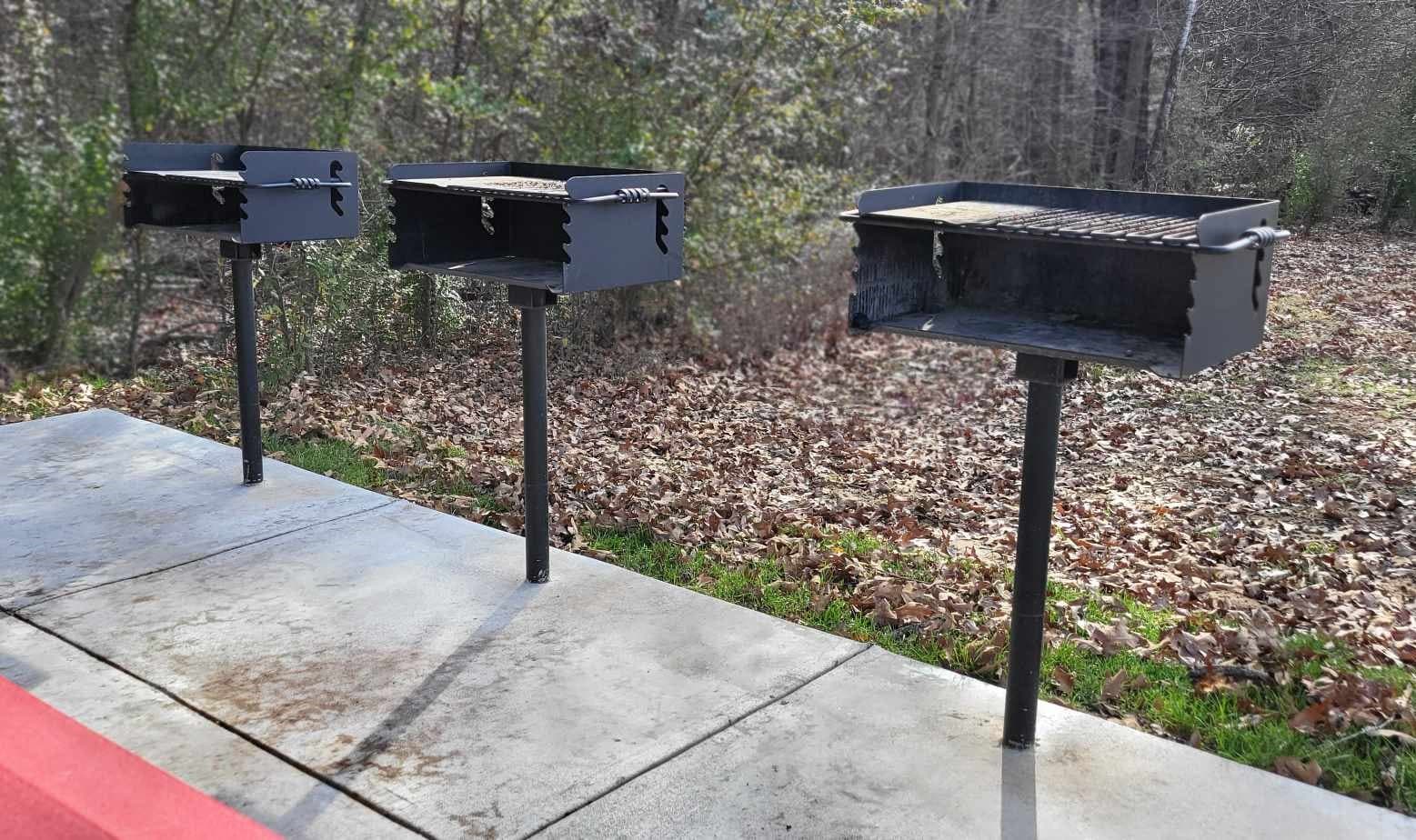 Three metal grills on pedestals, set against a backdrop of trees and fallen leaves.