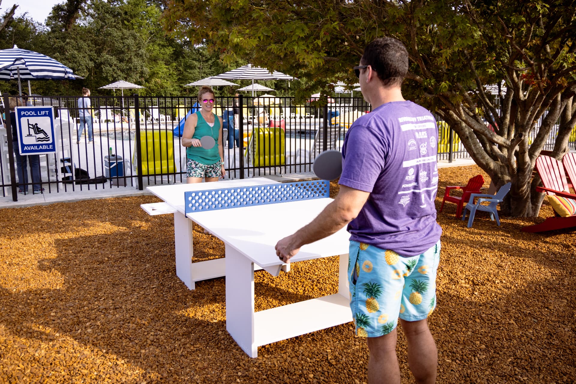 Two people are playing ping pong outdoors near a pool area.