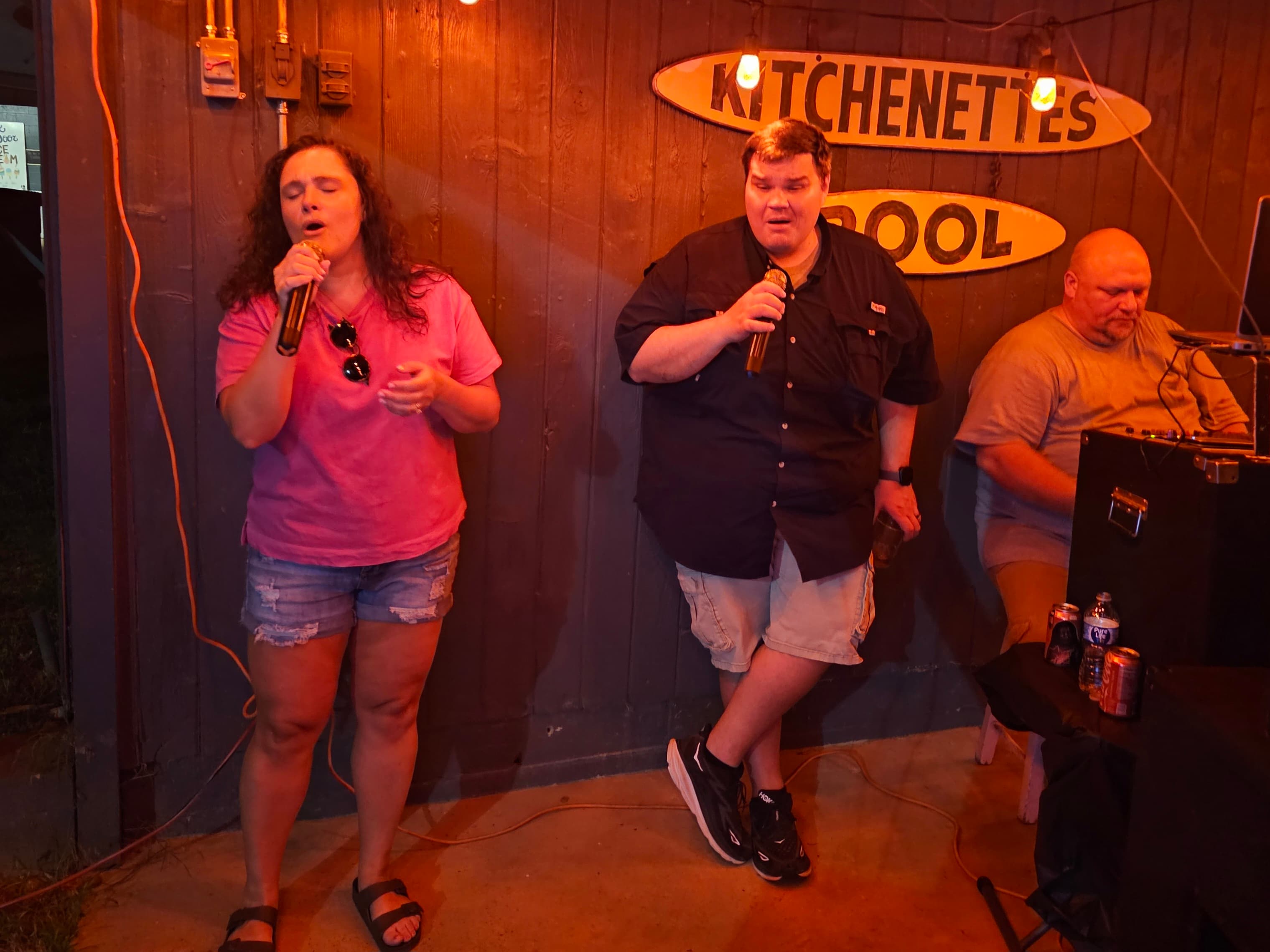 Two individuals sing karaoke while a third person plays music in the background at a casual outdoor setting.