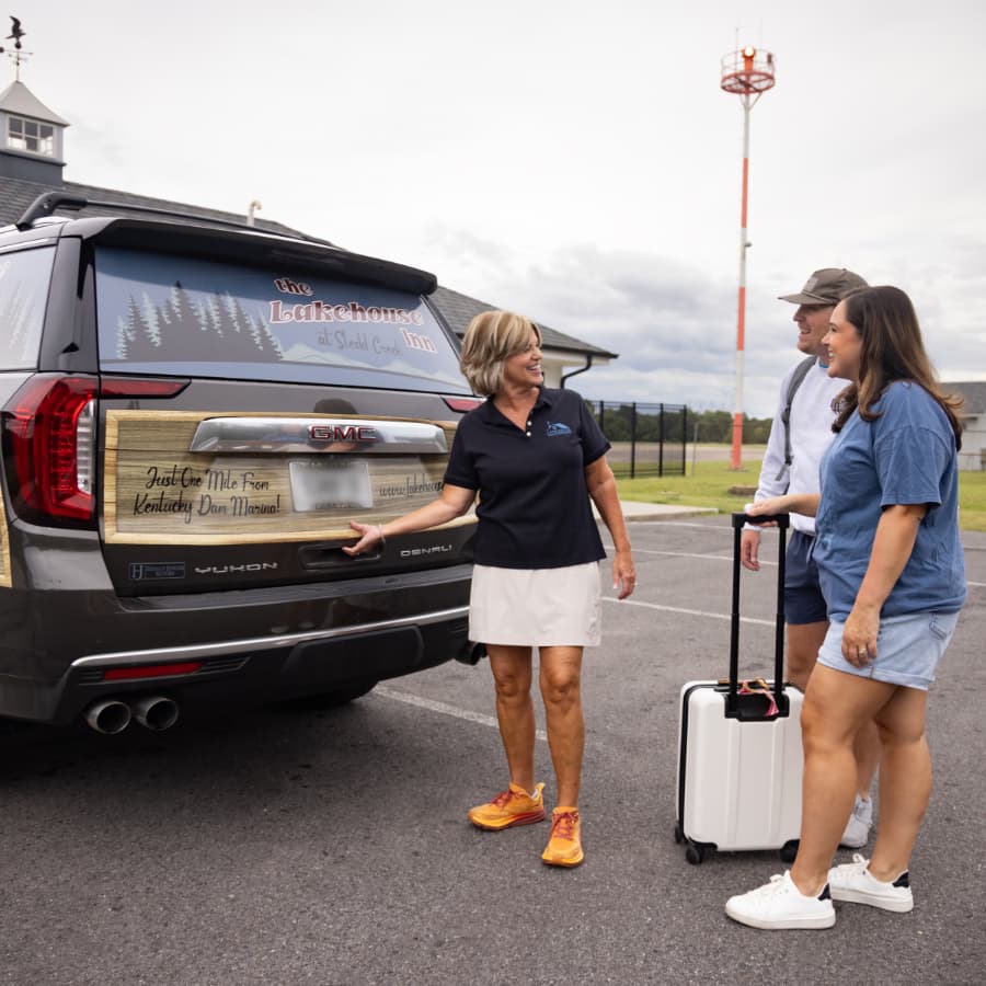 A woman in a polo shirt greets a couple at the back of a GMC SUV parked near a lake.