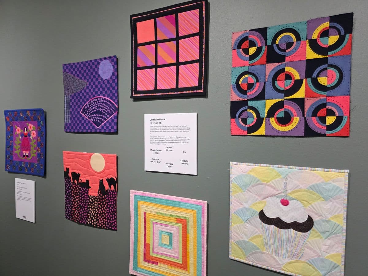 A display of colorful textile art pieces mounted on a gray wall, accompanied by descriptive labels.