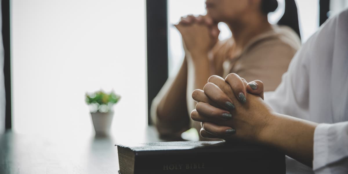 Two individuals are praying with hands clasped over a Bible, with a small potted plant in the background.