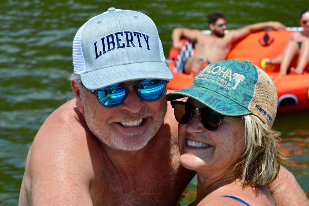 A smiling man and woman pose together in the water, wearing sunglasses and hats.