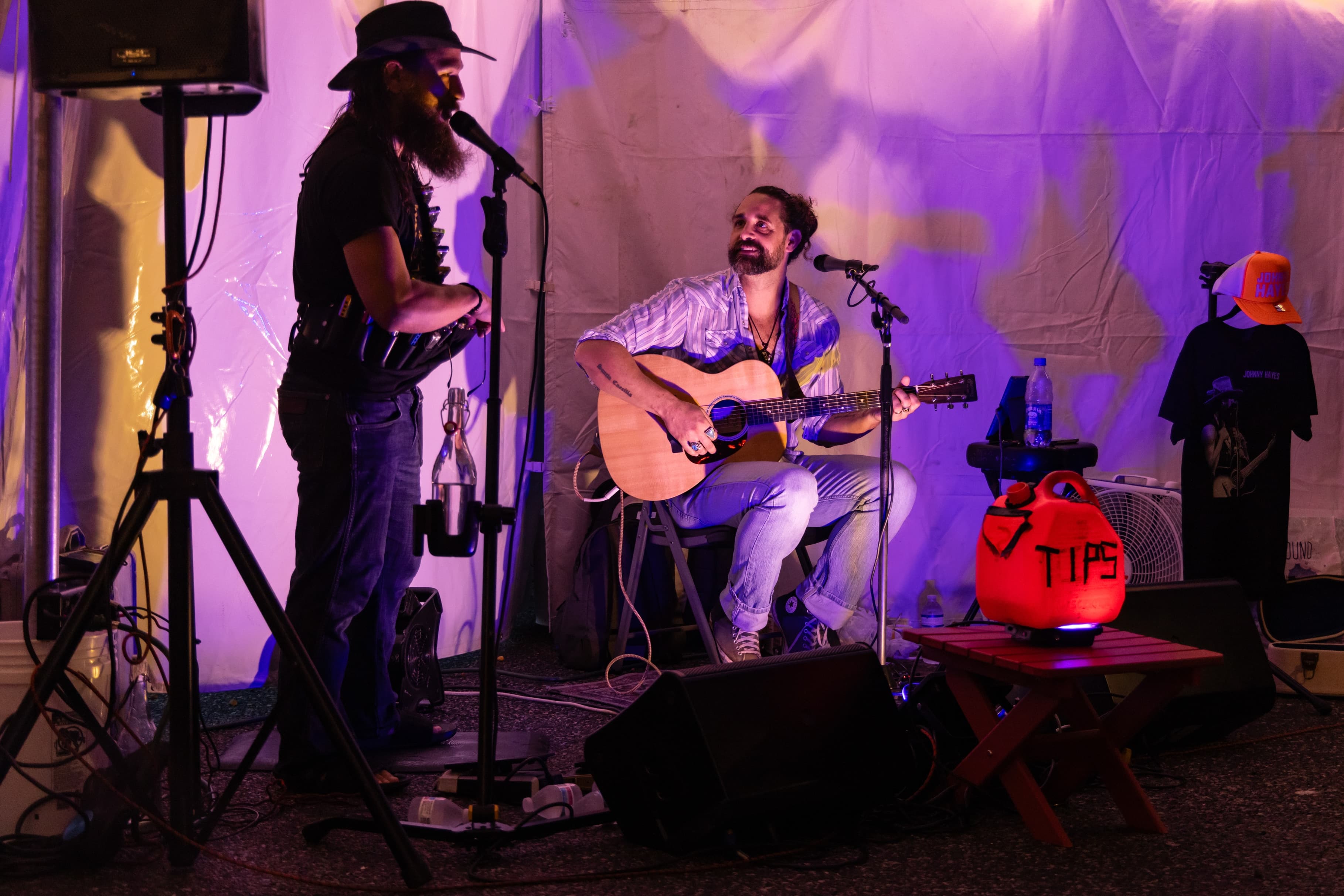 Two musicians perform on stage with a microphone and guitar, surrounded by colorful lighting and a tip jar.
