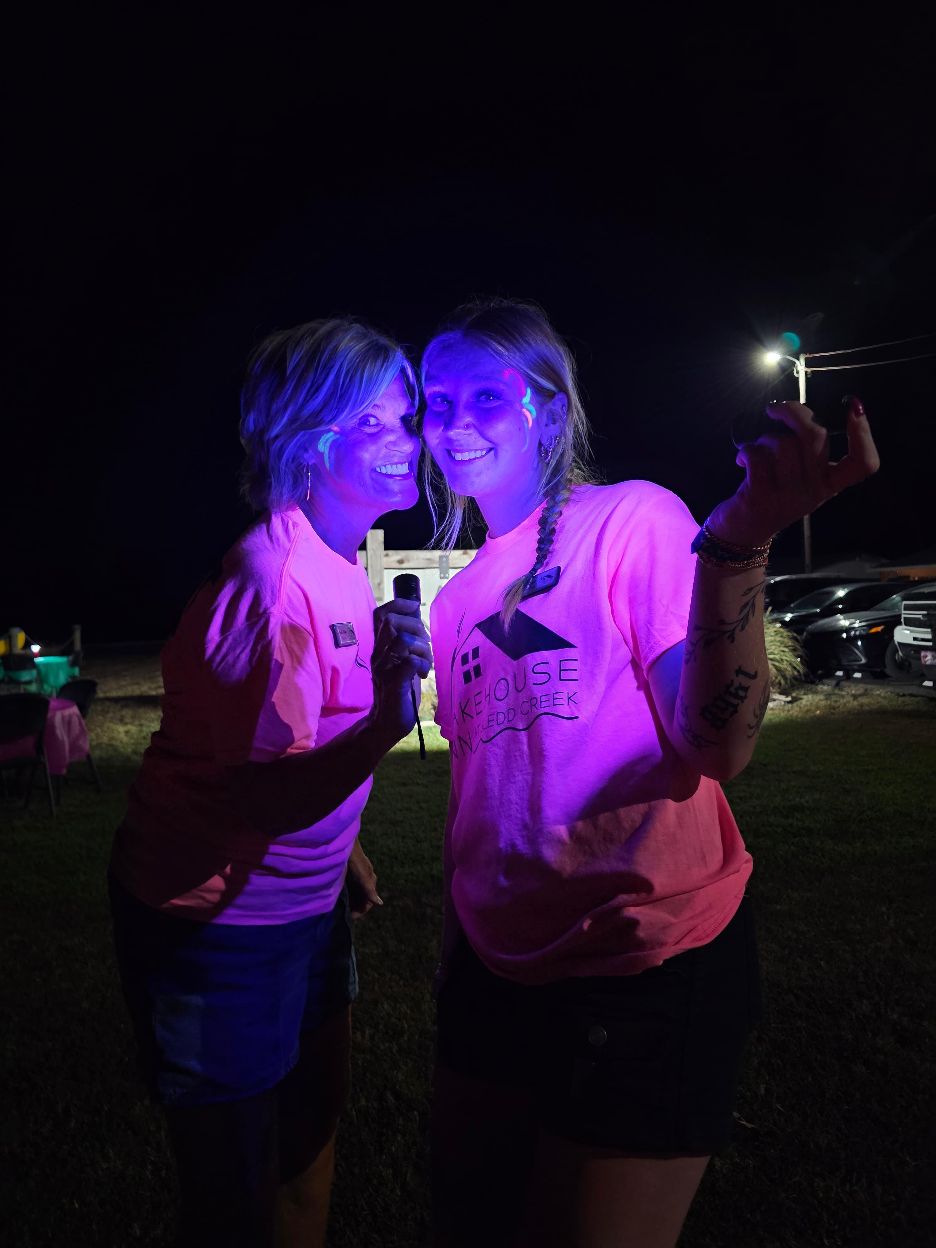 Two women wearing neon pink shirts pose playfully under blacklight at a night event.