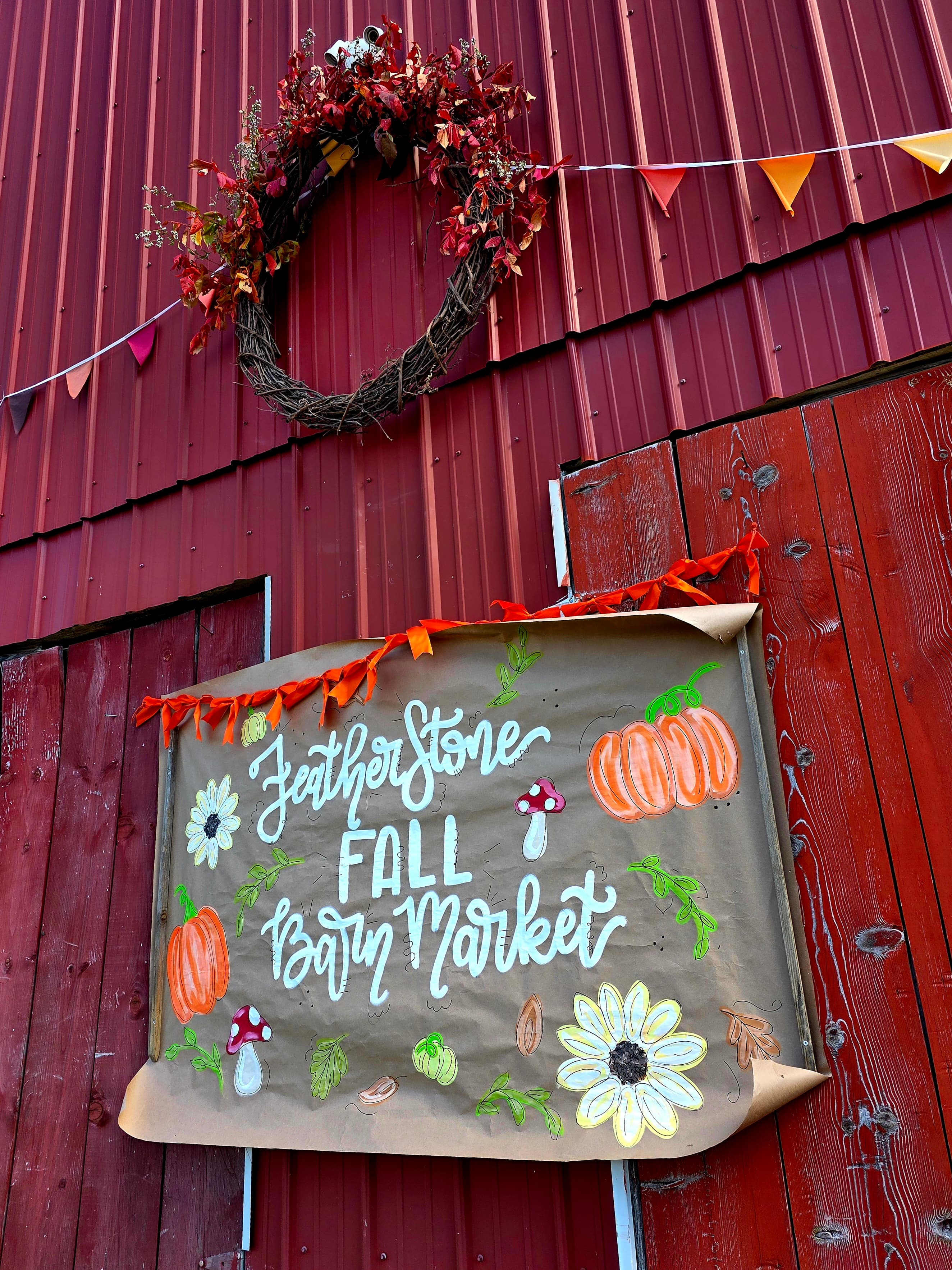 A decorated sign reading "Featherstone Fall Barn Market" with pumpkins and flowers hangs on a red barn.
