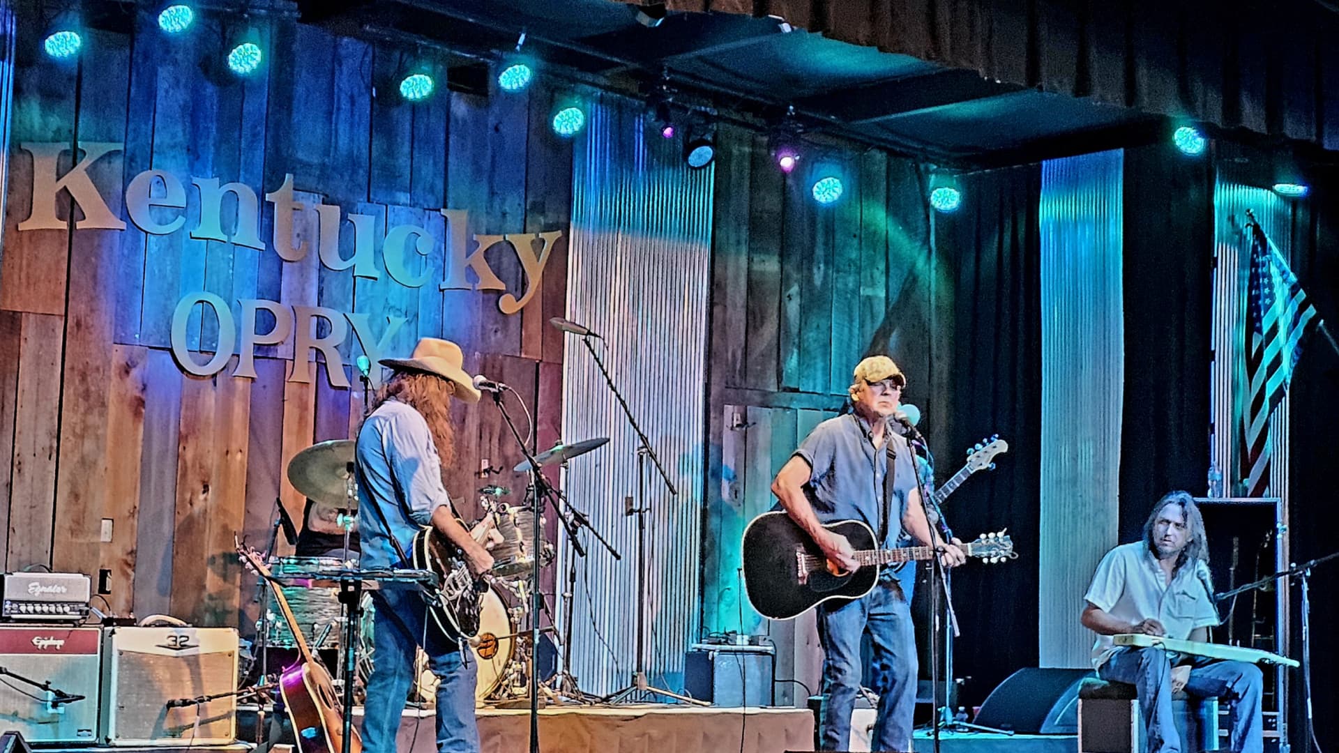 Three musicians perform on stage at the Kentucky Opry, with instruments and a wooden backdrop.
