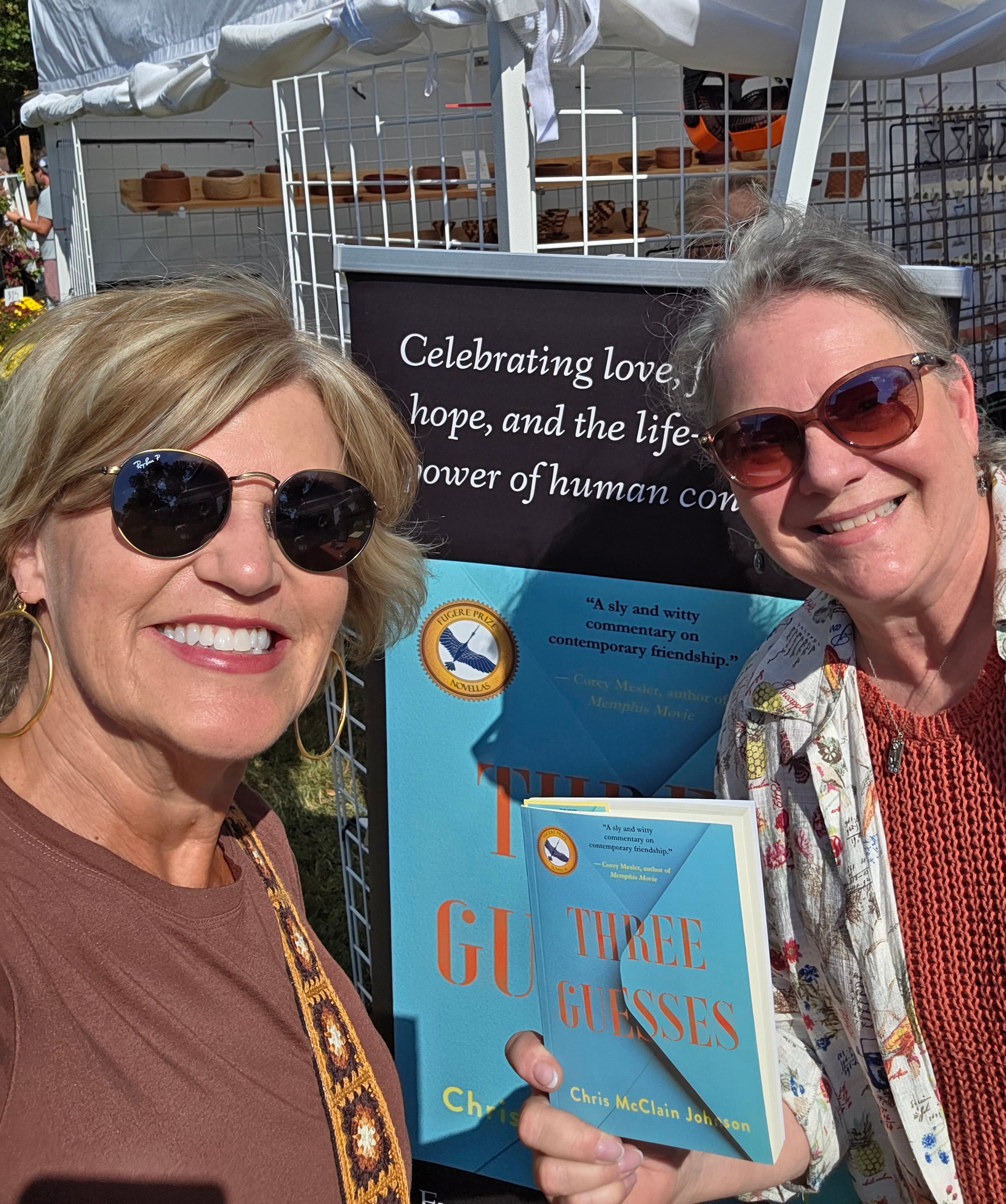 Two smiling women take a selfie holding the book titled "Three Guesses" at an outdoor event.