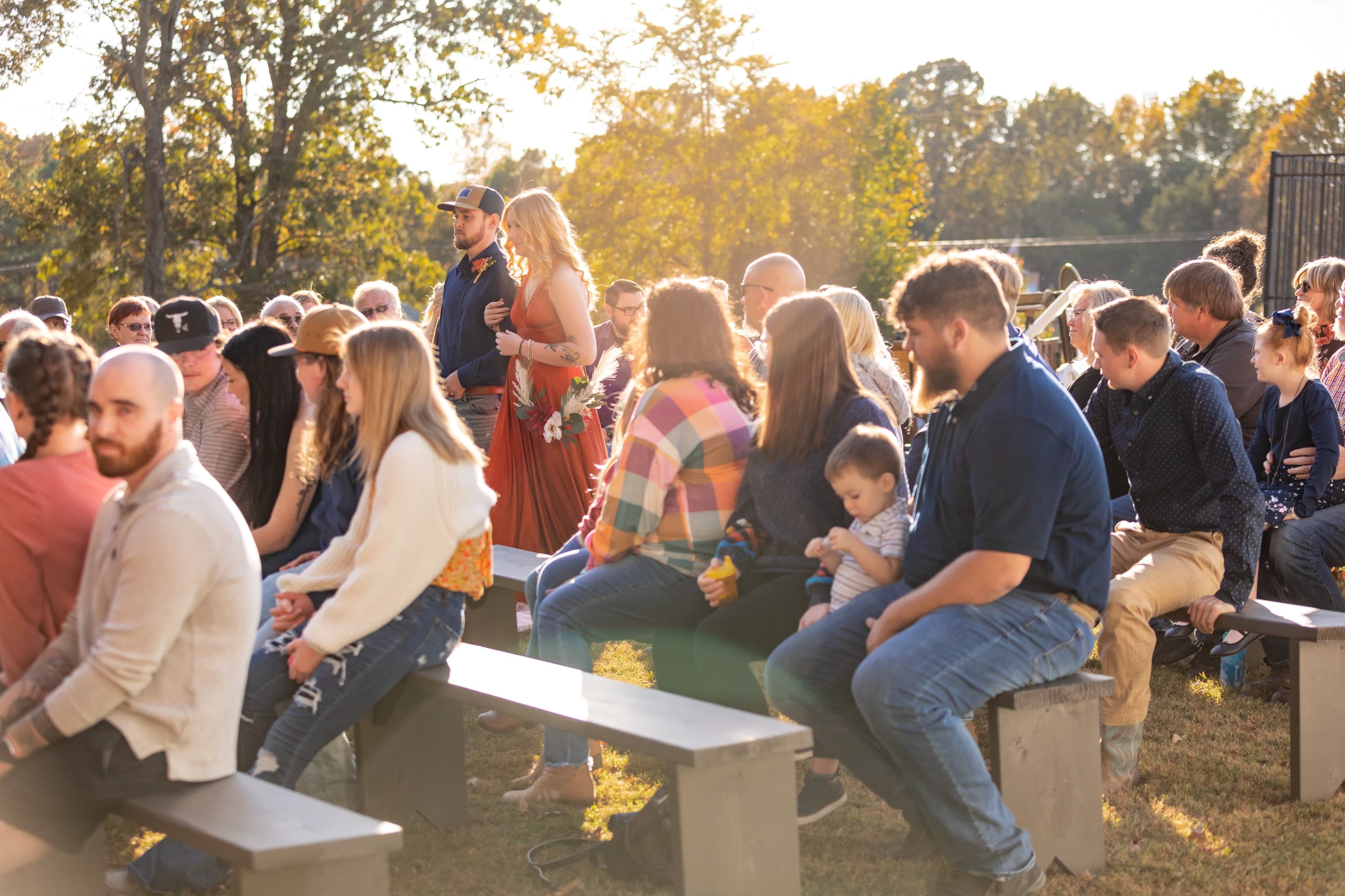 A couple walks down an aisle while seated guests watch in a sunlit outdoor setting.