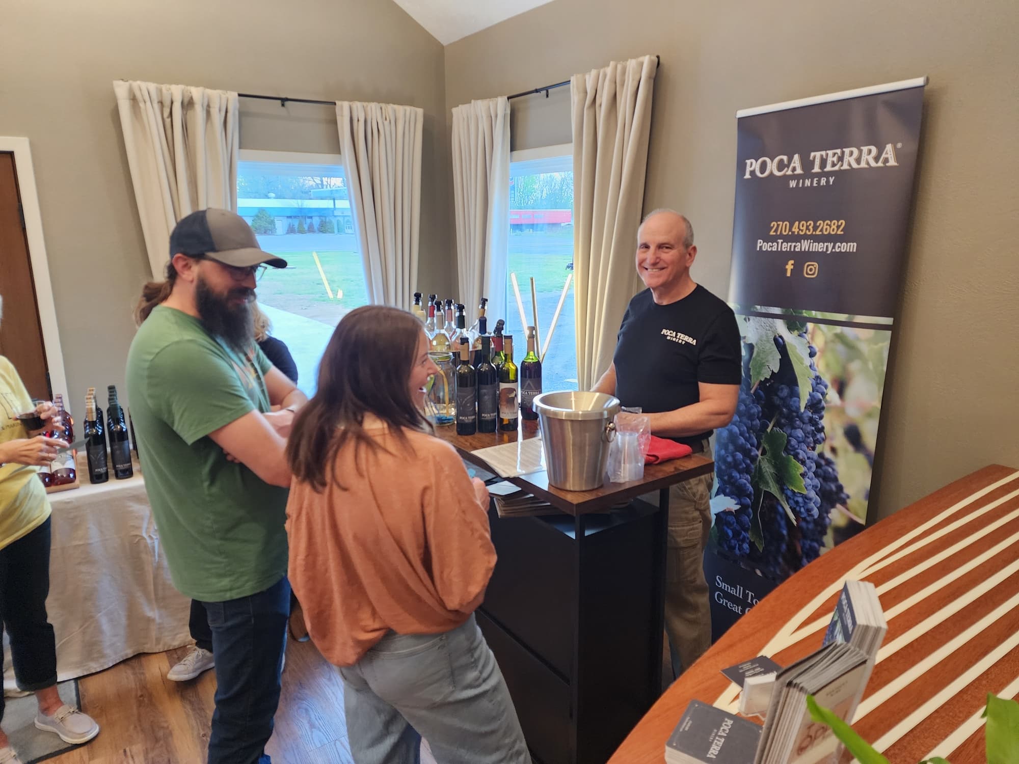 A group of people engage with a smiling winery representative at a tasting event featuring various wine bottles.