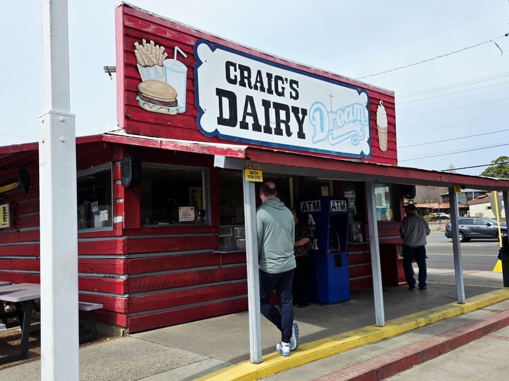 A small red wooden dairy stand with a sign reading "Craig's Dairy Dream" and customers at the counter. A small red wooden dairy stand with a sign reading "Craig's Dairy Dream" and customers at the counter.