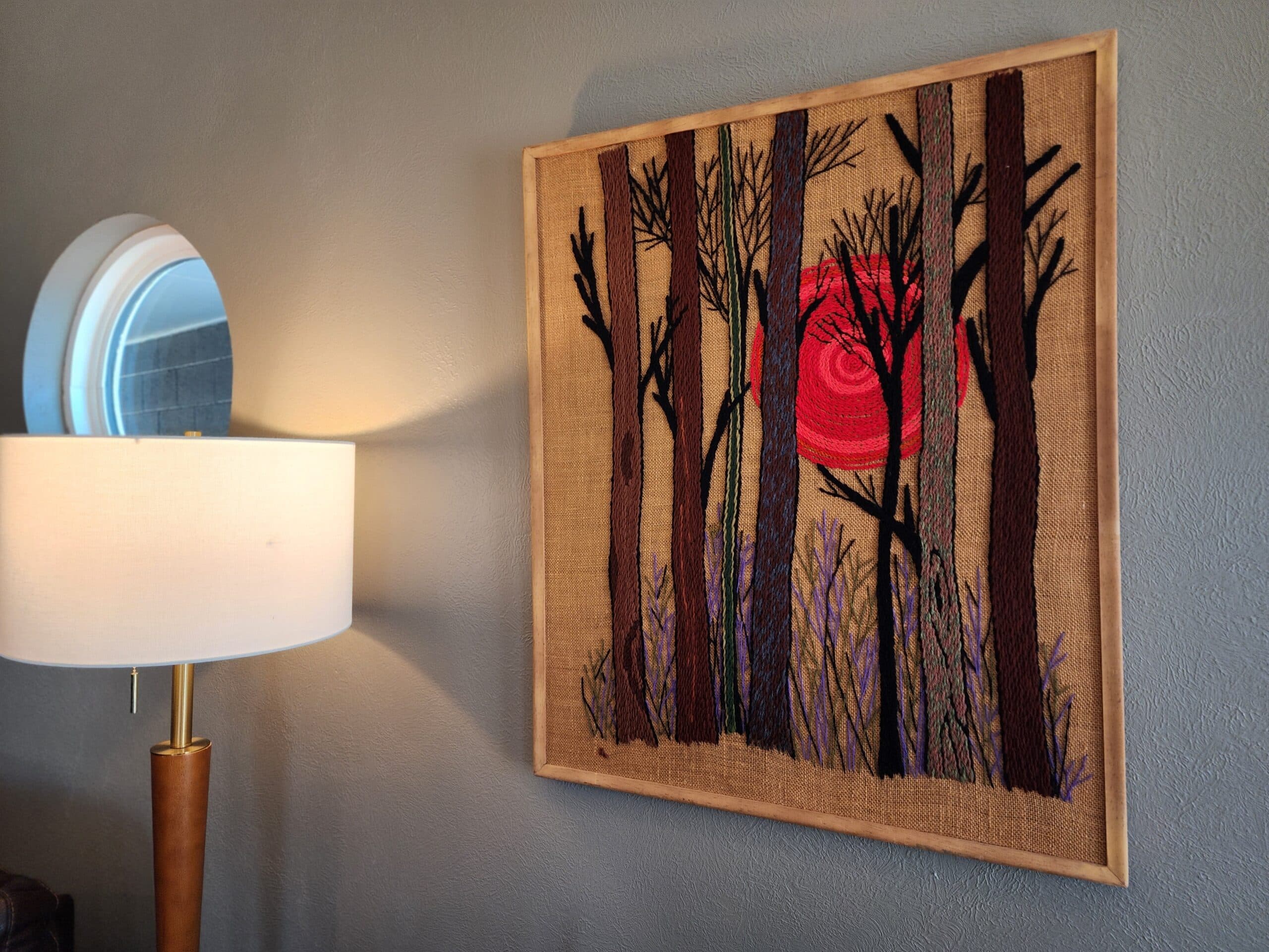 A framed textile artwork depicting silhouetted trees against a red circular background, with a lamp nearby. A framed textile artwork depicting silhouetted trees against a red circular background, with a lamp nearby.