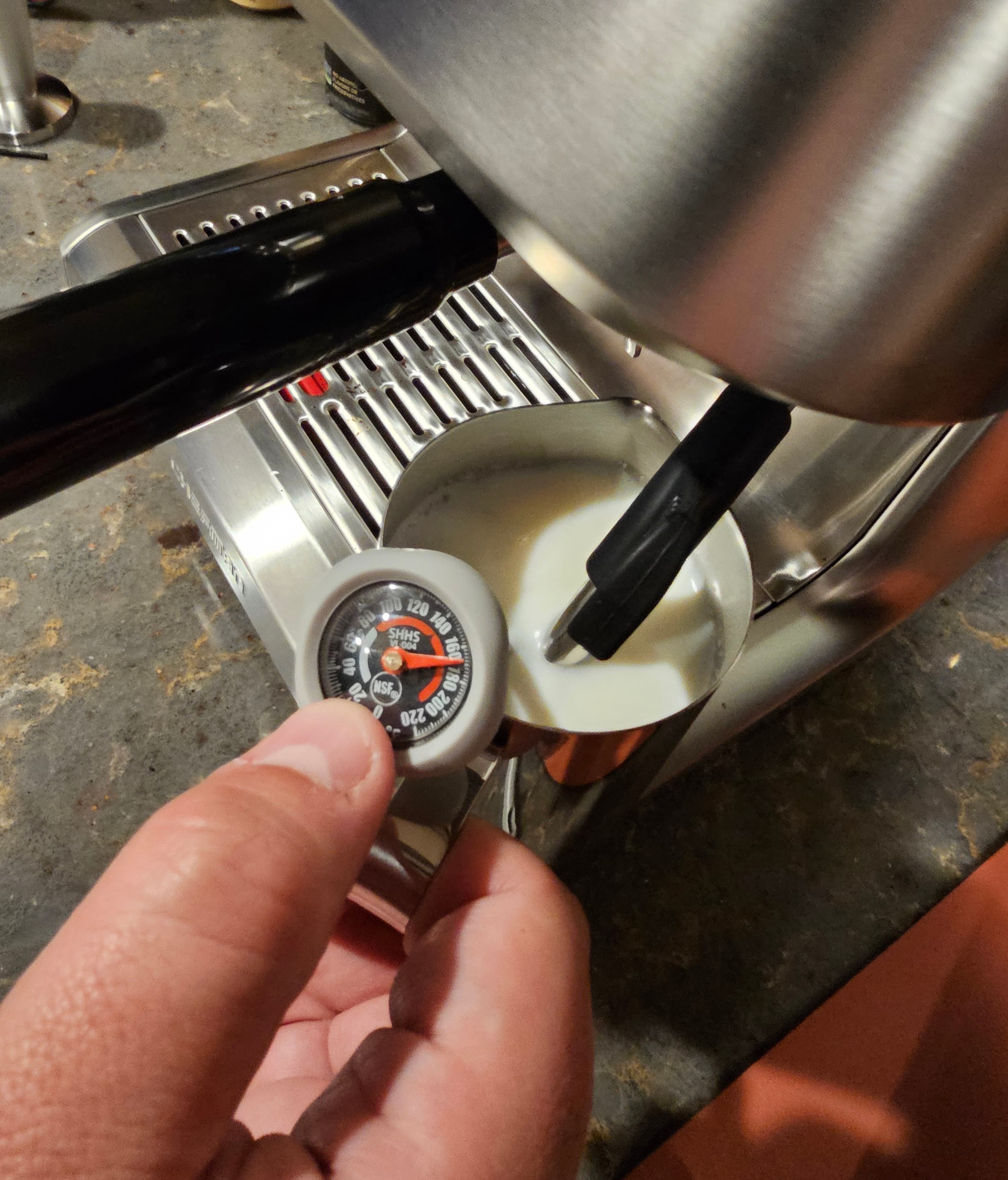 A hand holds a thermometer above a steaming milk pitcher next to an espresso machine.