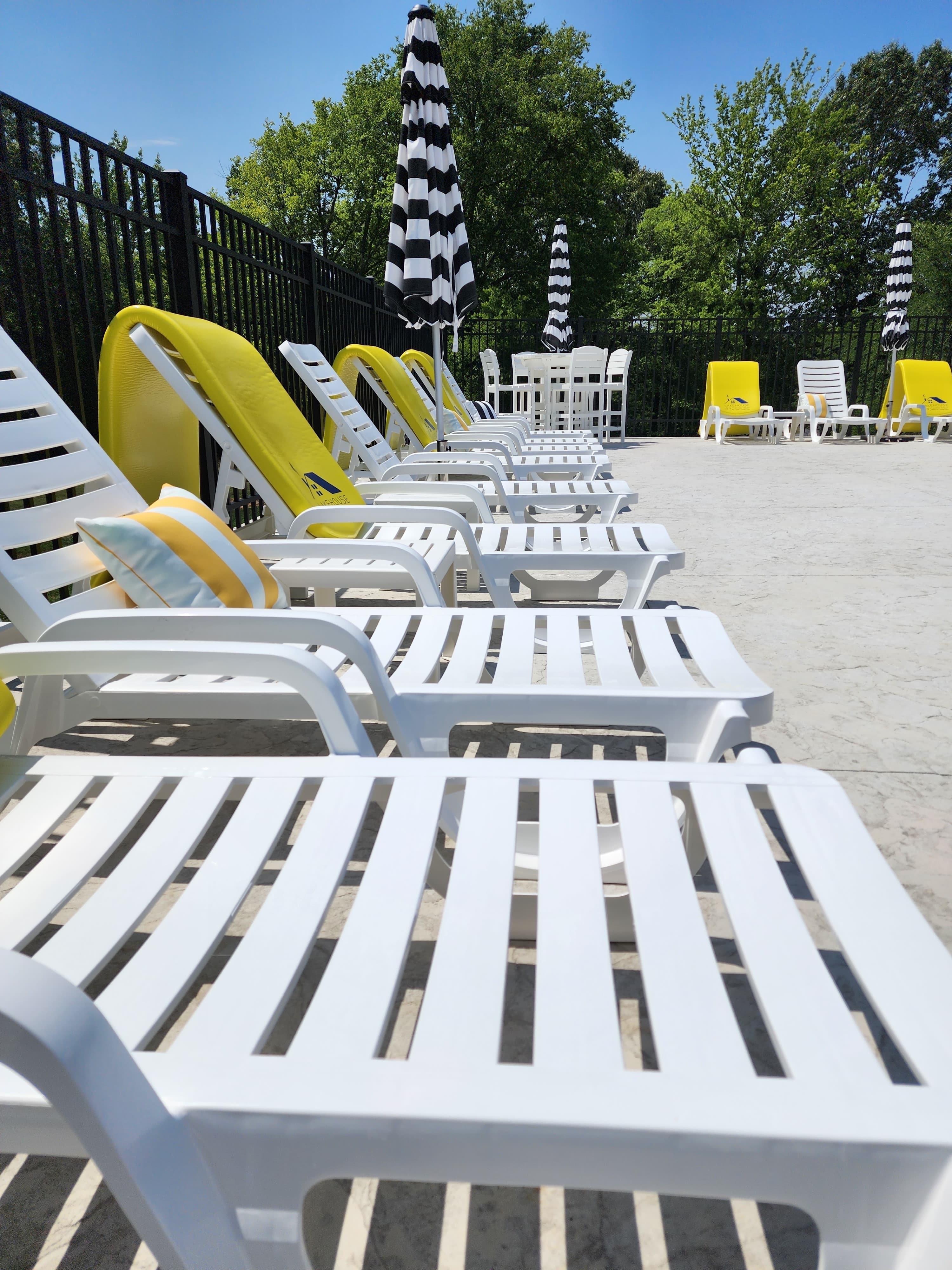 Row of white lounge chairs and yellow poolside loungers under striped umbrellas by a pool.