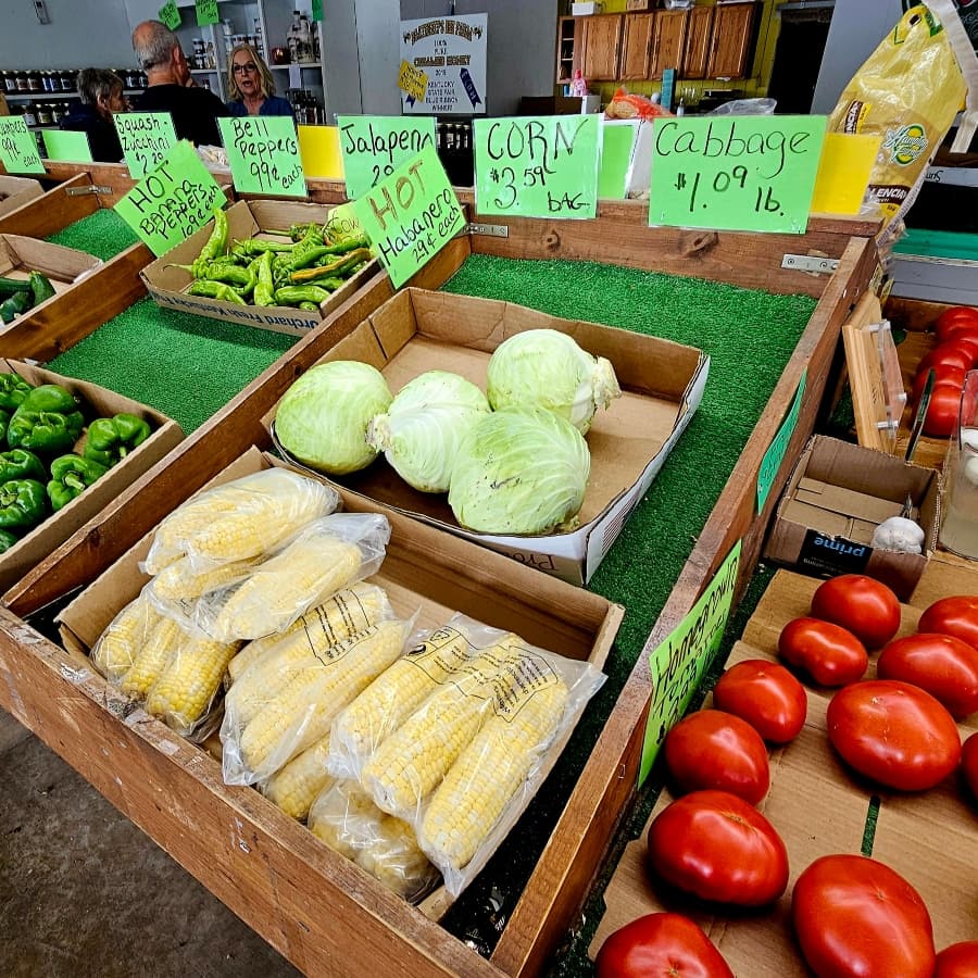 A variety of fresh produce, including corn, cabbage, jalapeños, and tomatoes, displayed in wooden crates at a farmer's market.
