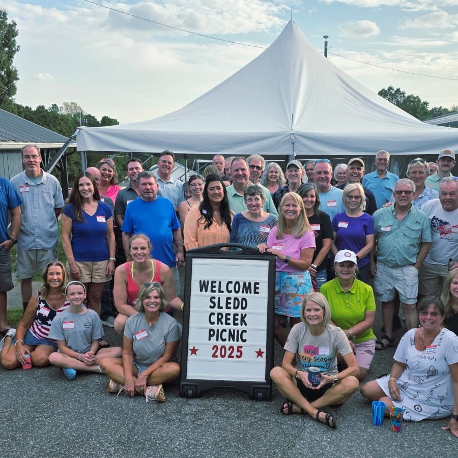 A large group of people gathers in front of a sign welcoming attendees to the Sledd Creek Picnic 2025 under a tent.
