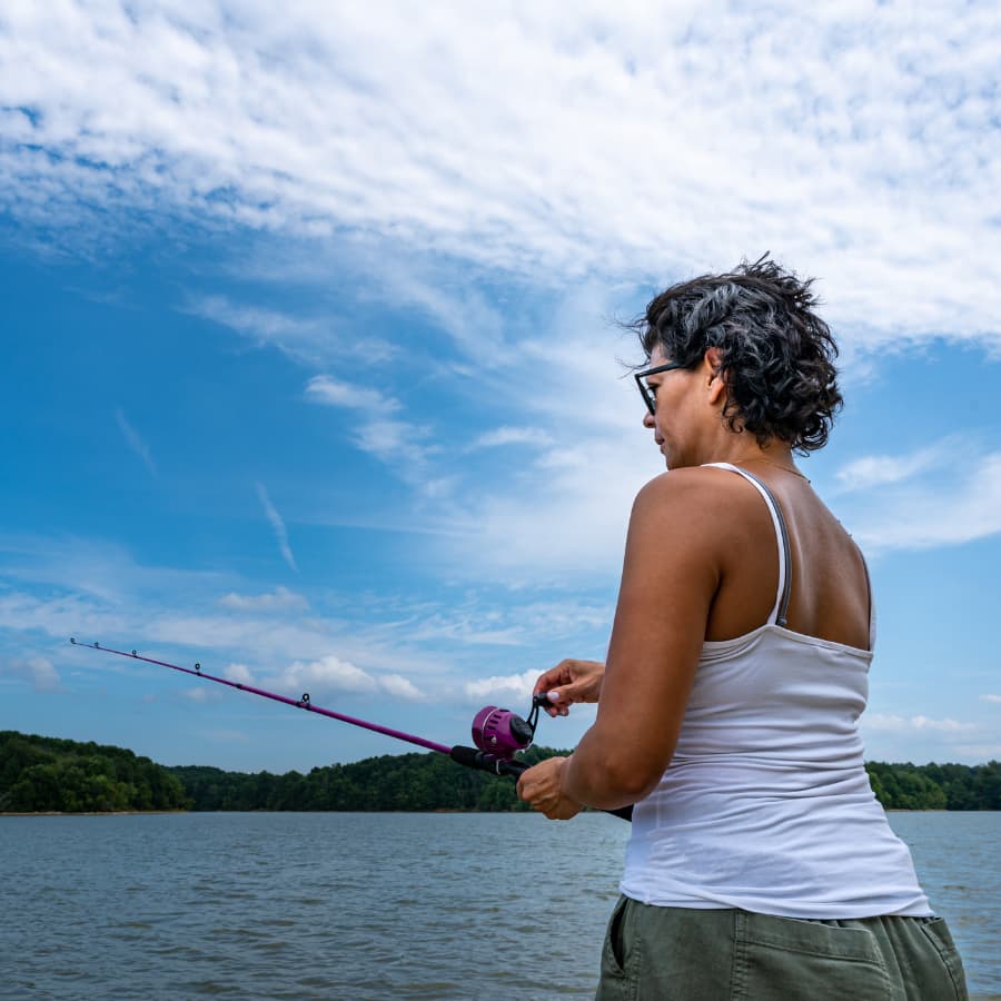 A woman in a tank top fishes from the shore under a blue sky.