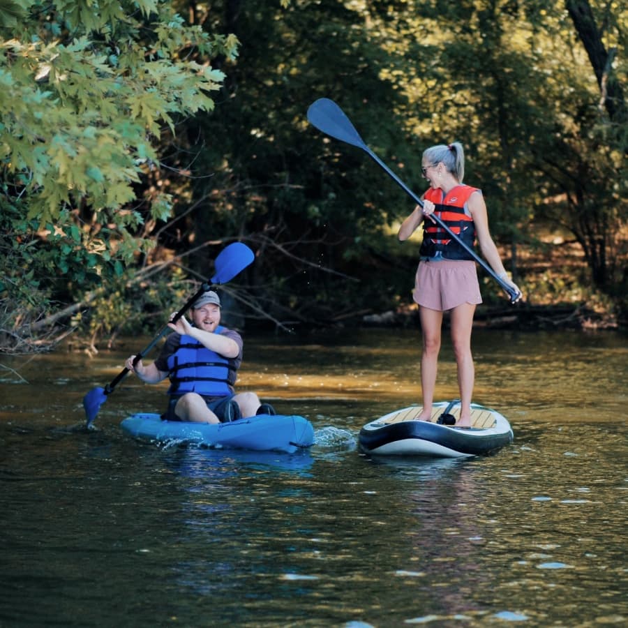 A woman stands on a paddleboard while a man paddles a kayak on a serene river.