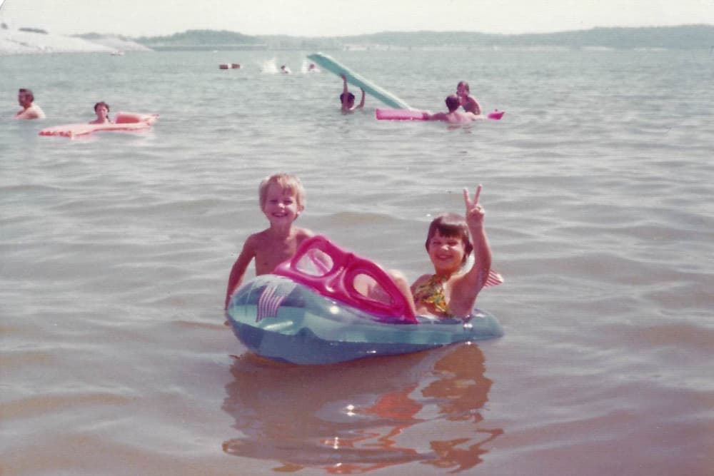Two children smiling in a small inflatable boat on a sunny day at the beach.