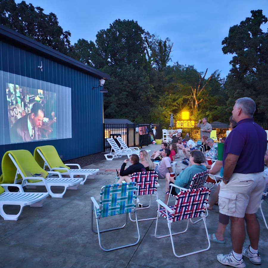A group of people watches a movie on an outdoor screen near a swimming pool at dusk.