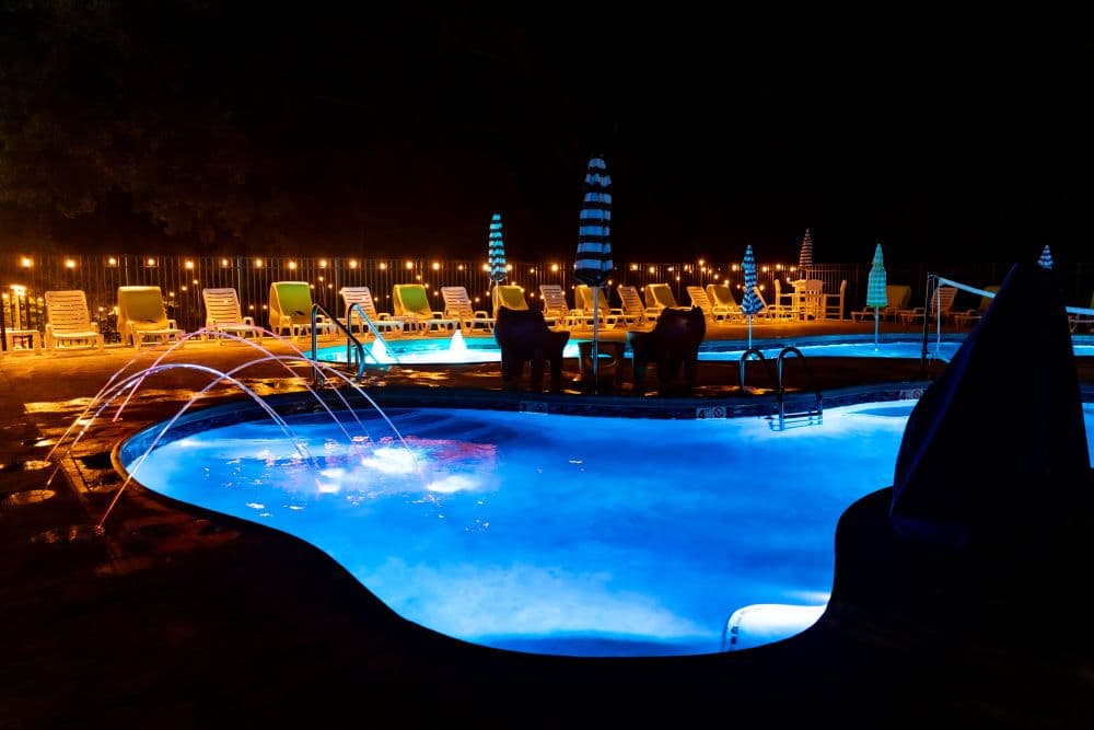 A brightly lit swimming pool at night with colorful lights and lounge chairs around it.