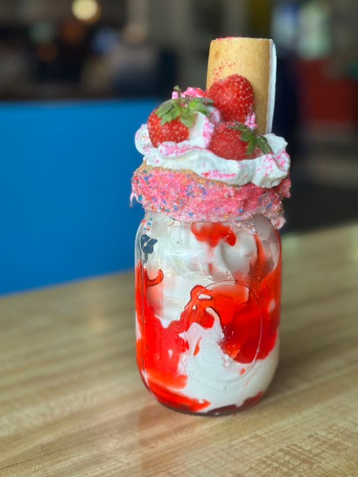 A decorative milkshake in a mason jar topped with whipped cream, strawberries, and colorful sprinkles.