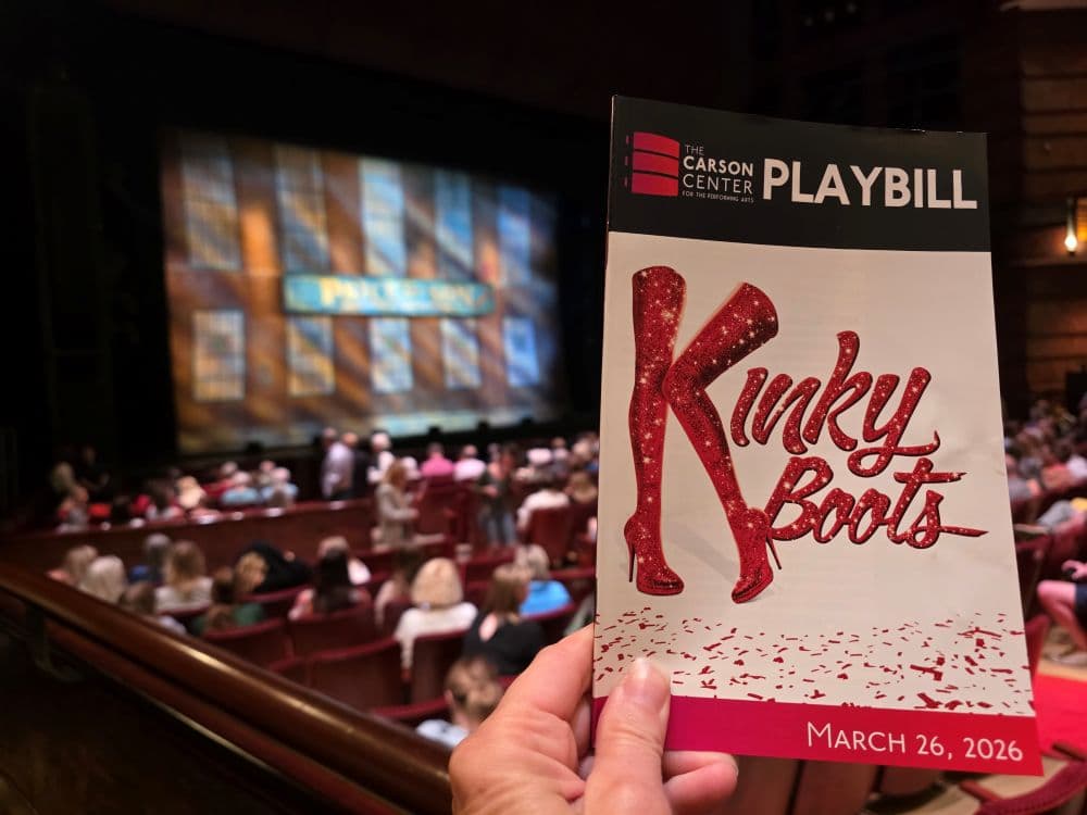 A hand holds a Playbill for the musical "Kinky Boots" in a theater with an audience and stage backdrop visible. A hand holds a Playbill for the musical "Kinky Boots" in a theater with an audience and stage backdrop visible.