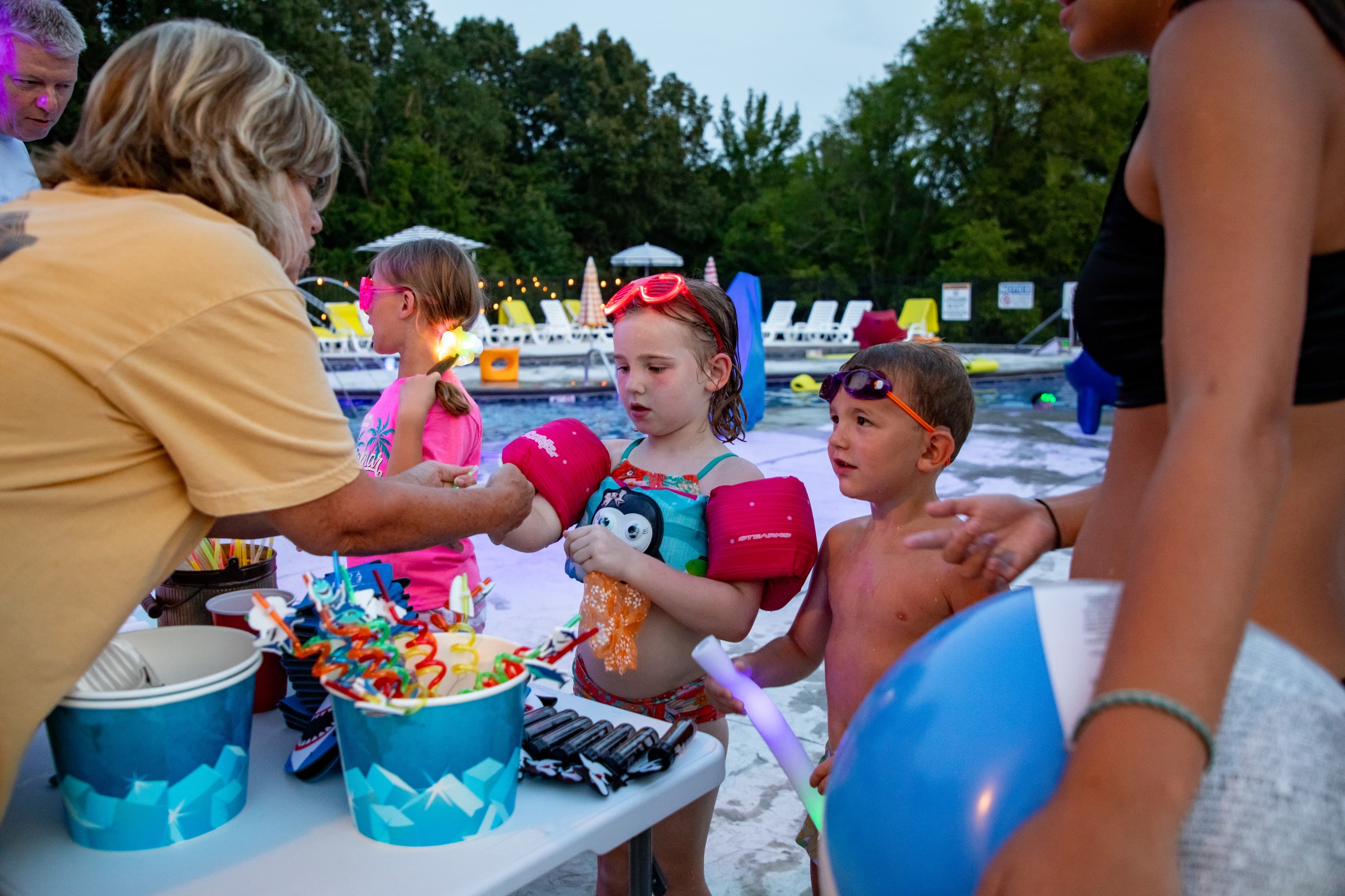 Children enjoying a nighttime pool party while receiving colorful treats from an adult.