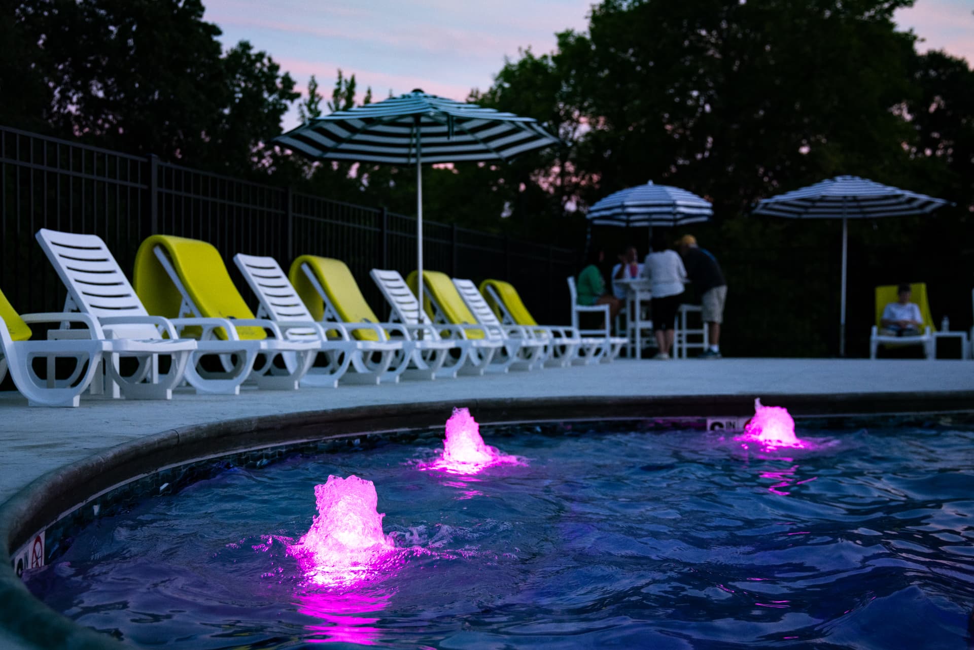 A pool with pink fountains and yellow lounge chairs is lined with umbrellas as people gather nearby.