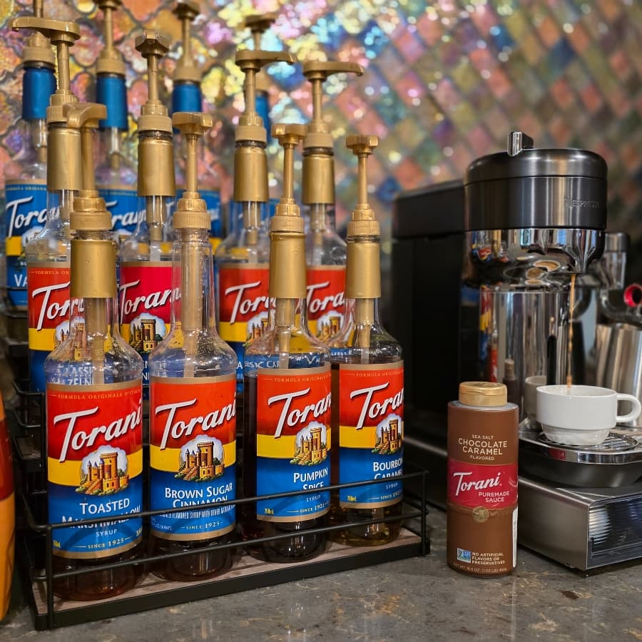 A collection of Torani syrup bottles displayed on a shelf next to an espresso machine and a cup.