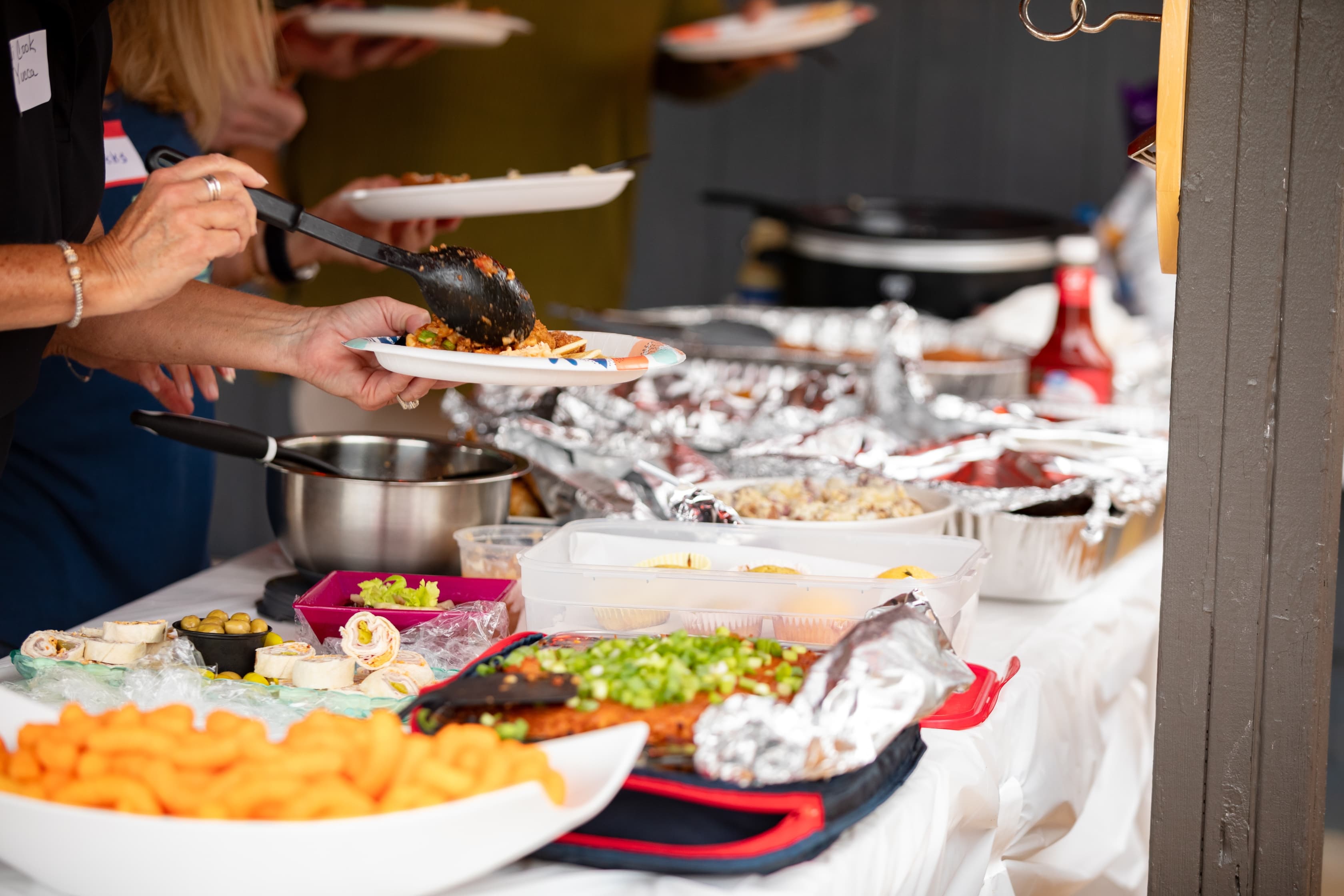 A close-up of hands serving food from a buffet table filled with various dishes.