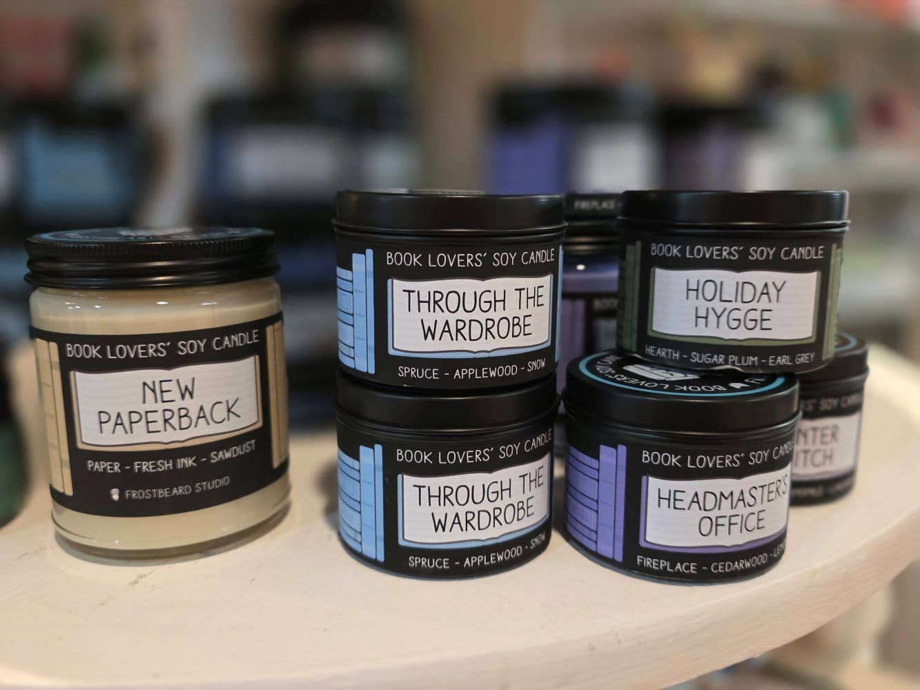 A collection of soy candles with various scents, including "New Paperback" and "Holiday Hygge," displayed on a shelf.