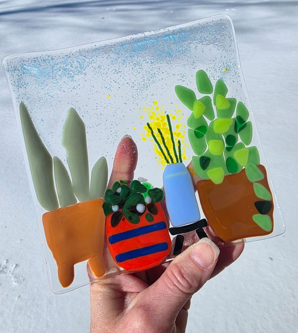 A hand holds a colorful glass art piece featuring various stylized potted plants.