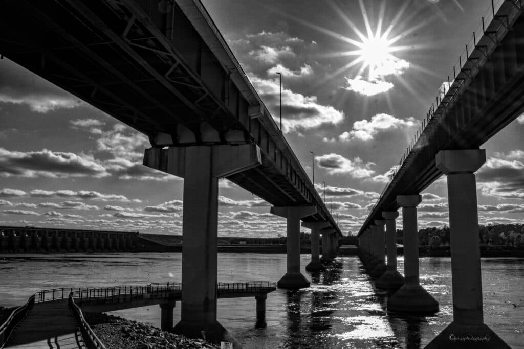 Black and white image of two bridges arching over a river, with sun rays shining through clouds above.