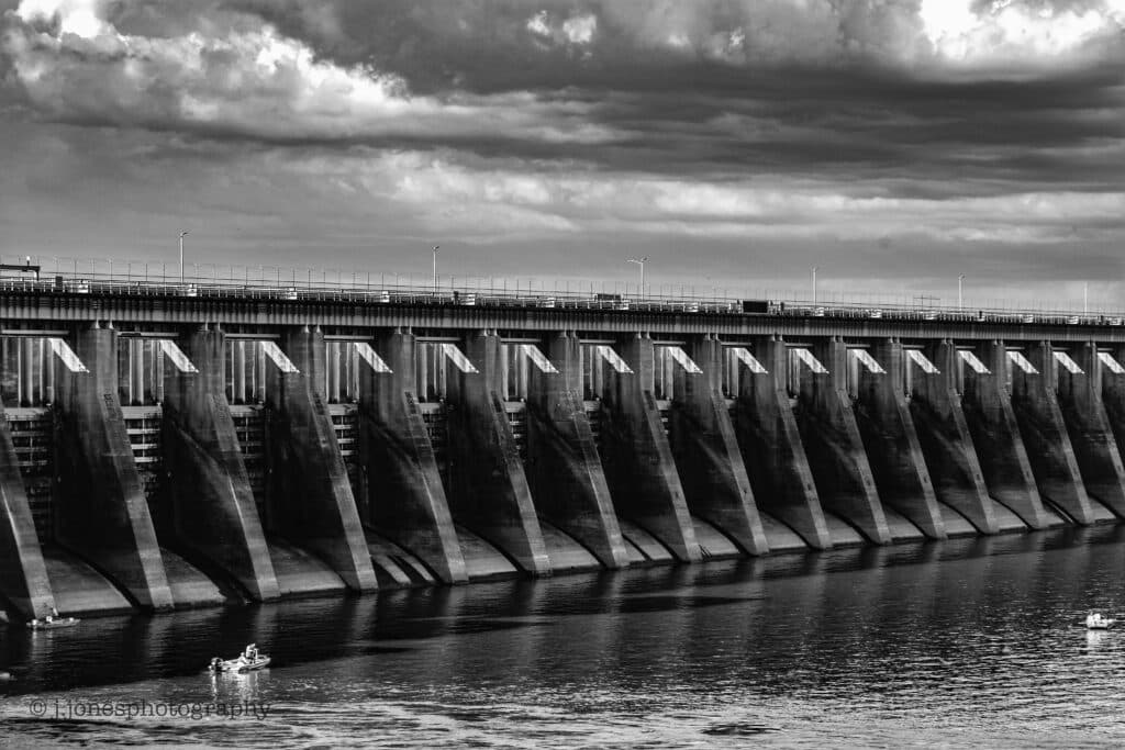 A black and white image of a large dam reflecting in the water, with clouds overhead.