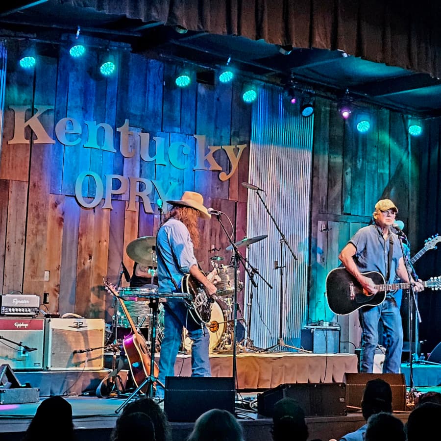 Two musicians perform on stage at the Kentucky Opry surrounded by musical instruments and colorful stage lights.