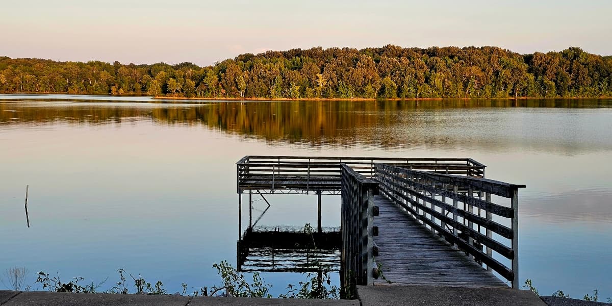 A serene view of a wooden dock extending into a calm, reflective lake surrounded by trees.