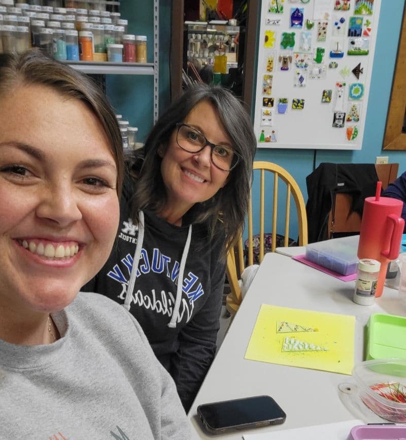 Two smiling women pose for a selfie at a craft table with art supplies.