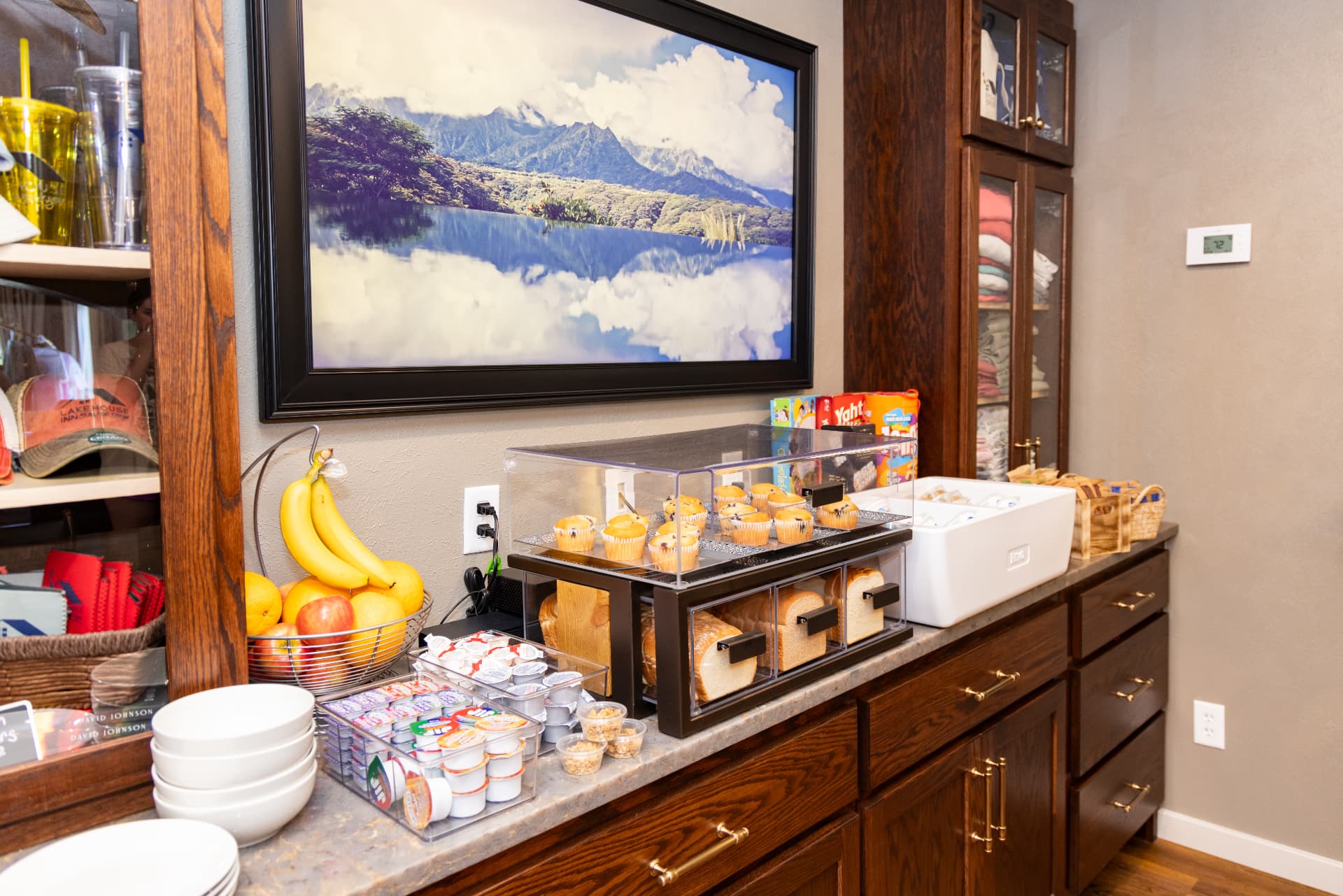 A kitchen countertop display features muffins, fruits, condiments, and a scenic mountain photograph.