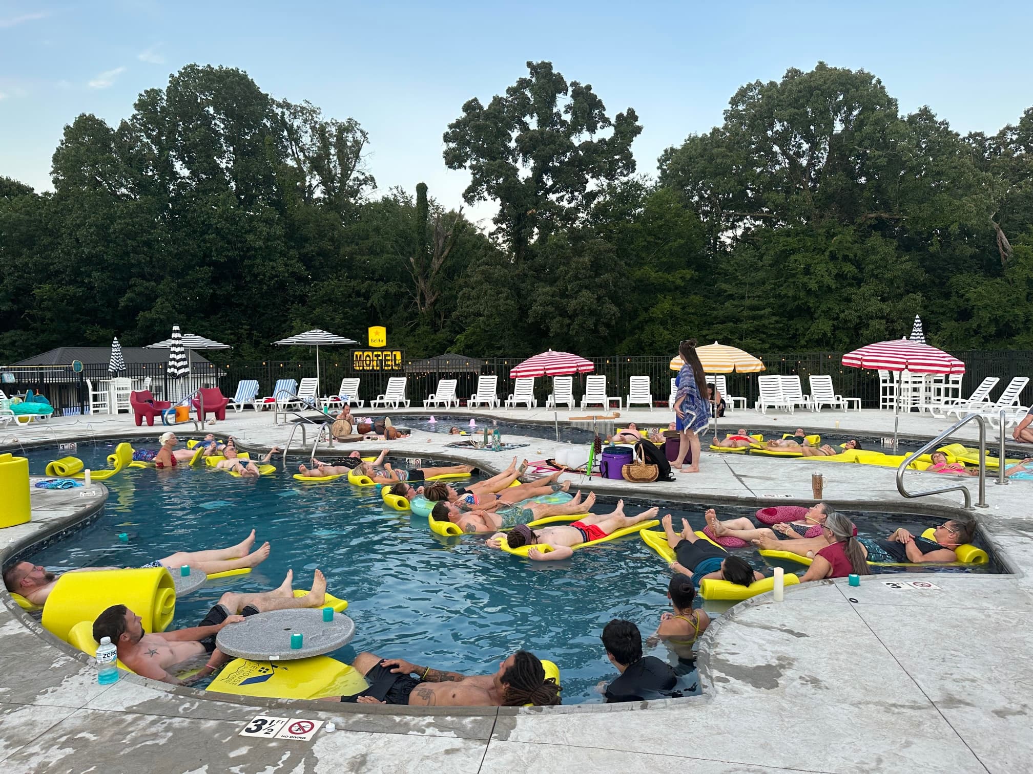 A crowded swimming pool with people lounging on yellow floats under colorful umbrellas.