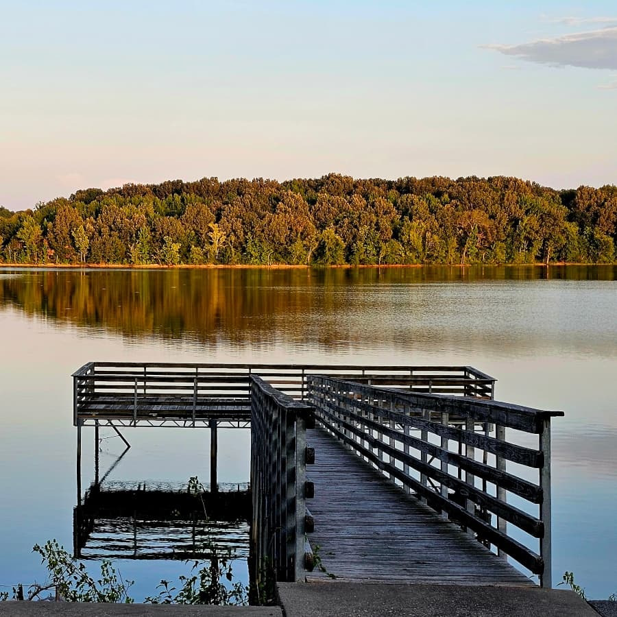 A wooden dock extends over a calm lake, reflecting the surrounding trees at sunset.