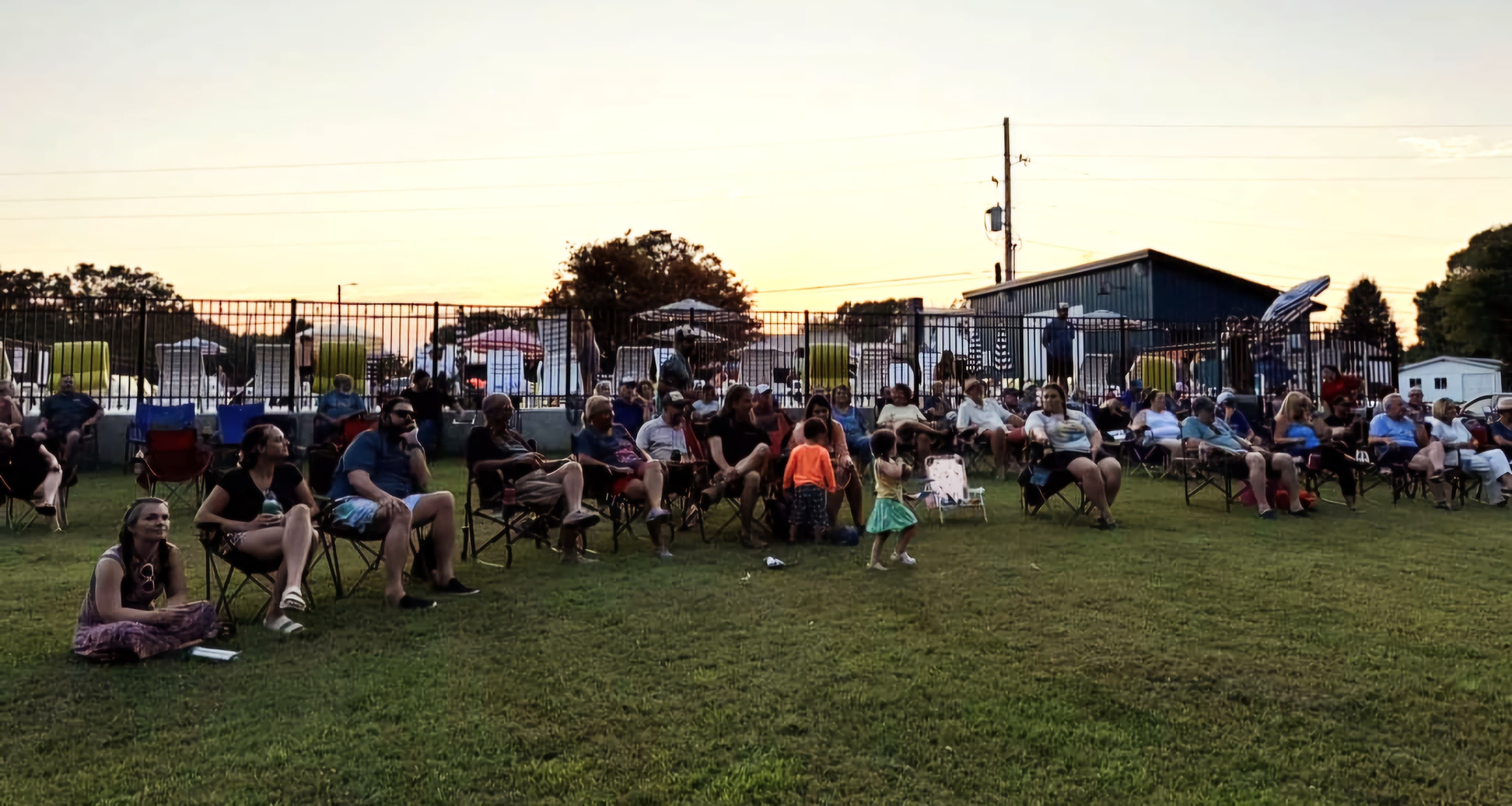 A crowd of people sits on lawn chairs enjoying an outdoor event as the sun sets.