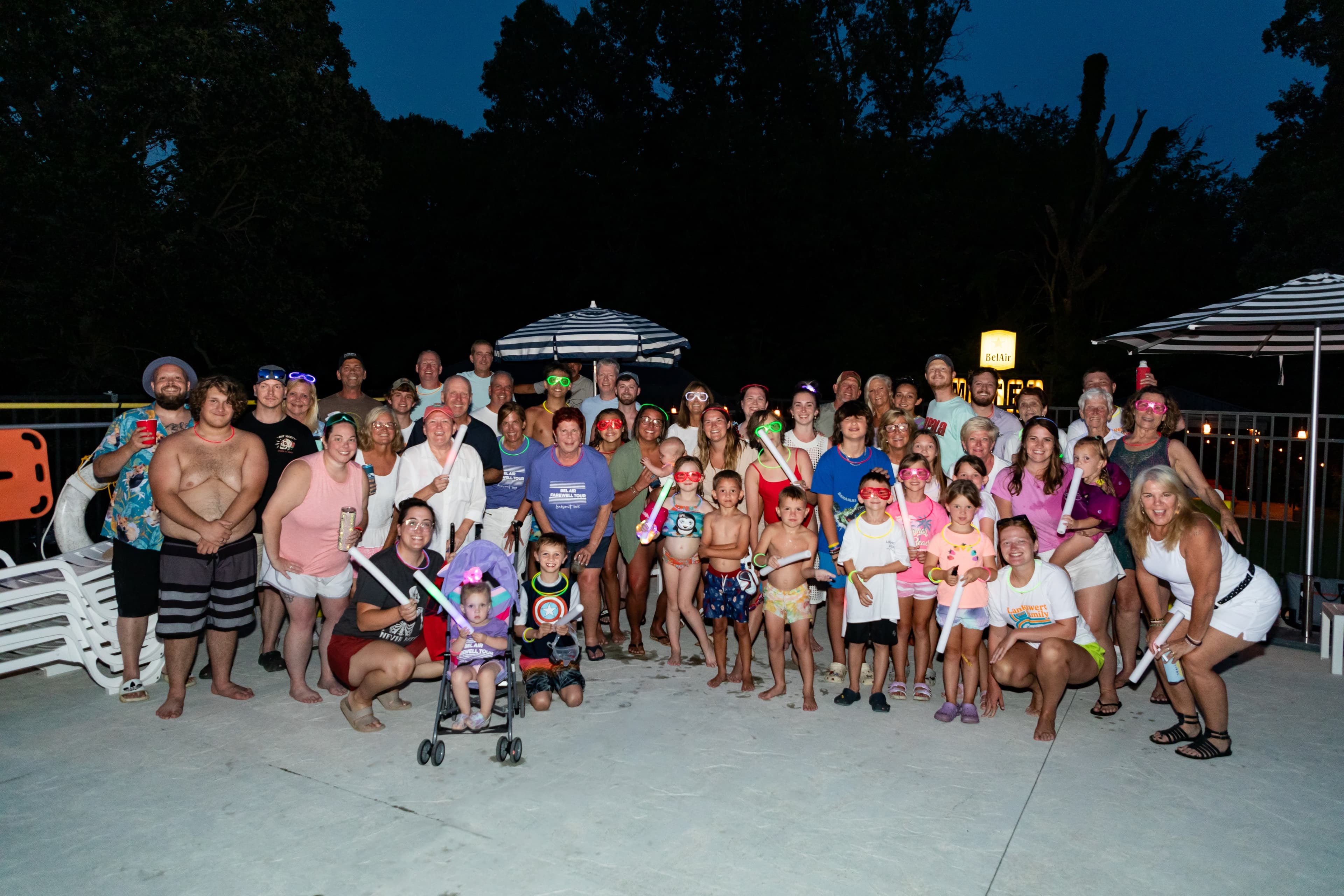 A large group of people, including children and adults, gather by a pool at night, all smiling and holding colorful light sticks.