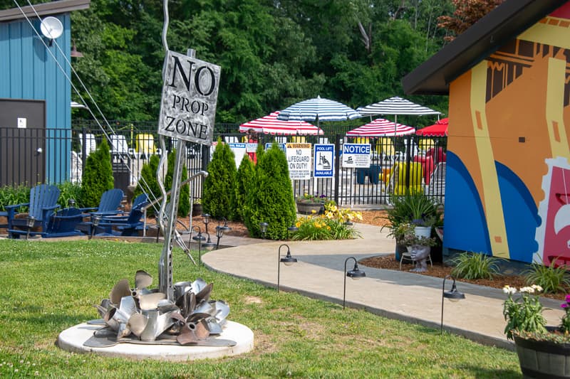 A decorative sign saying "NO PROP ZONE" stands in a lush green area near colorful umbrellas and lounge chairs. A decorative sign saying "NO PROP ZONE" stands in a lush green area near colorful umbrellas and lounge chairs.