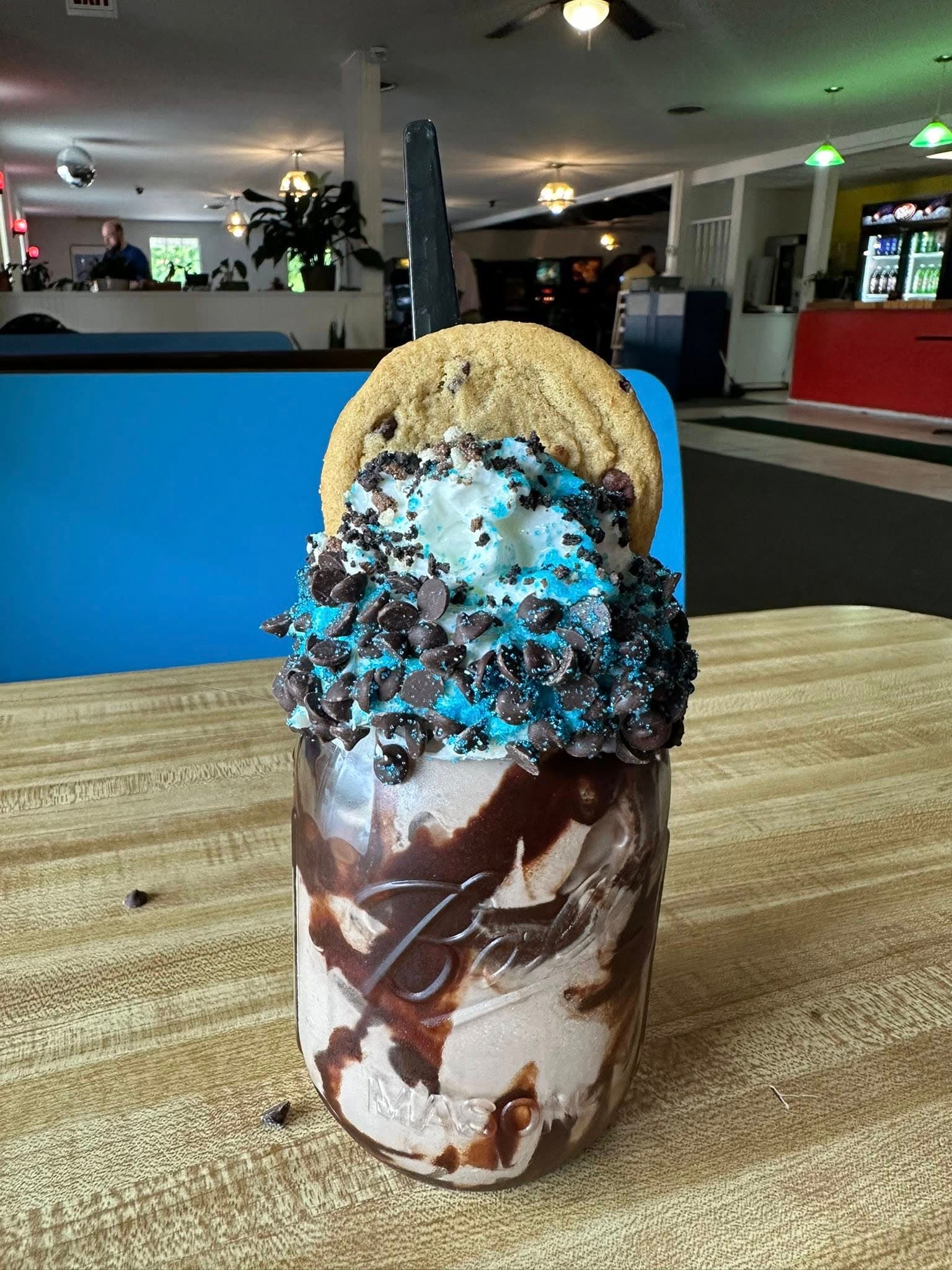 A decadent dessert featuring a jar filled with chocolate ice cream, topped with whipped cream, crushed cookies, blue sprinkles, and a cookie on top.