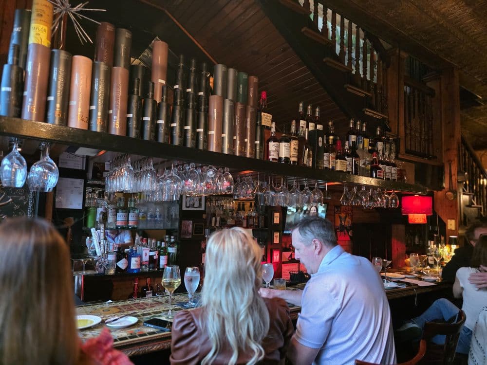 A rustic bar scene with bottles and glasses neatly arranged on shelves, and patrons seated at the counter.