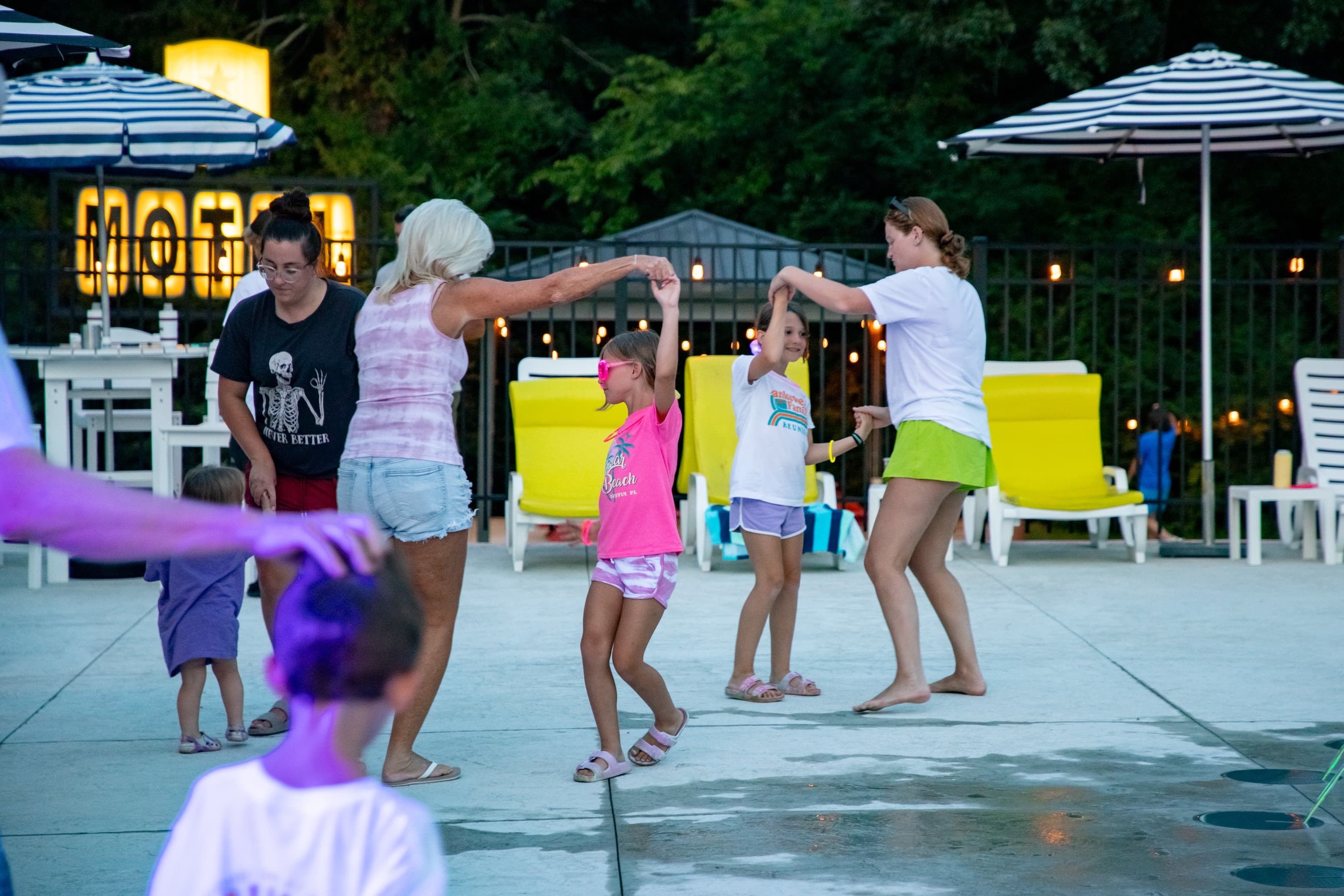 A group of women and children enjoy dancing by a poolside in the evening.