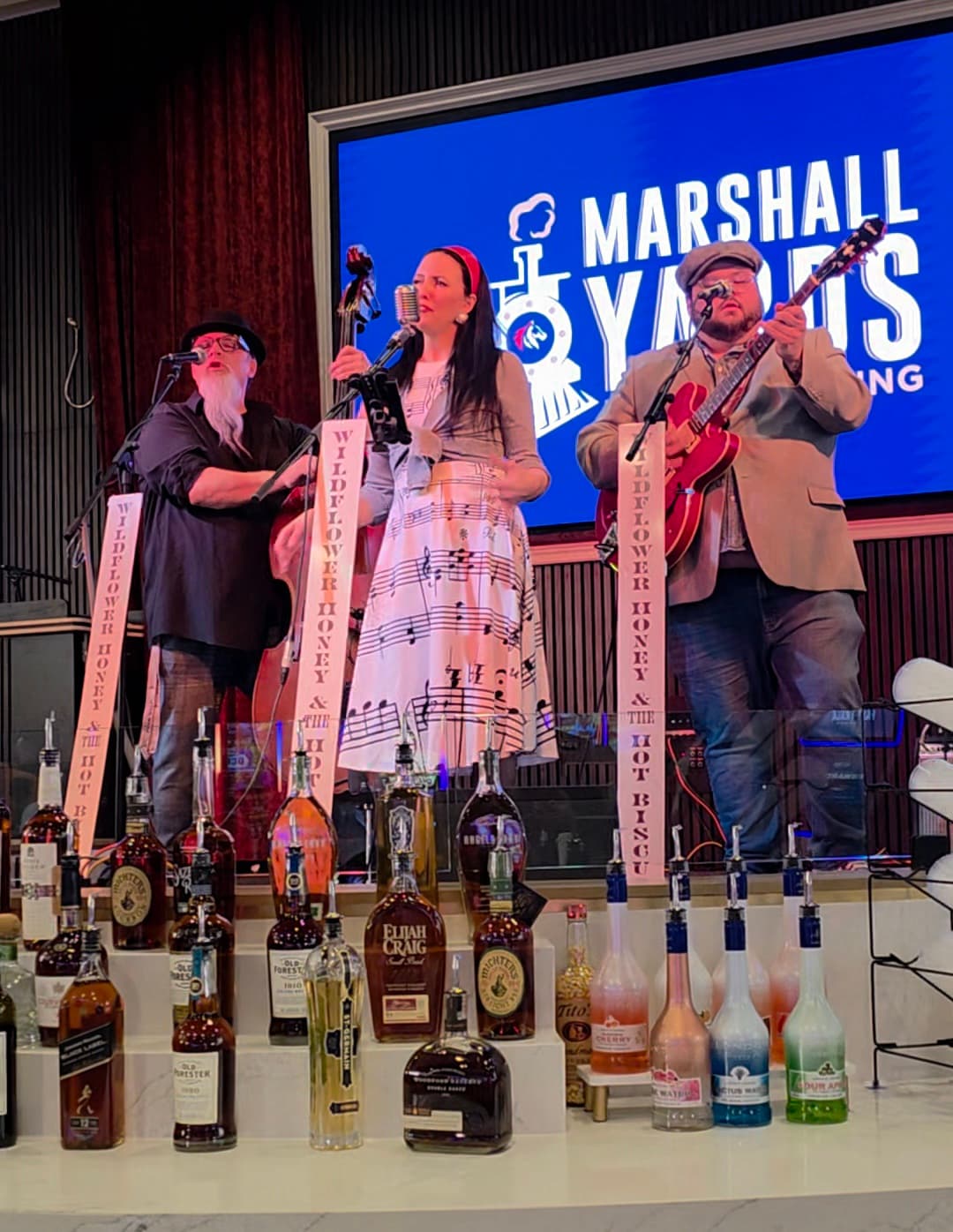 Three musicians perform on stage in front of a large screen, with a variety of liquor bottles displayed in the foreground.
